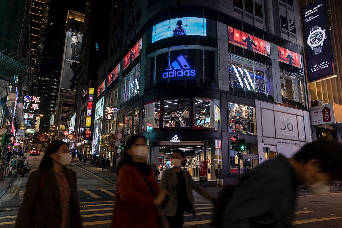 Pedestrians wearing face masks walk past the Adidas store and logo