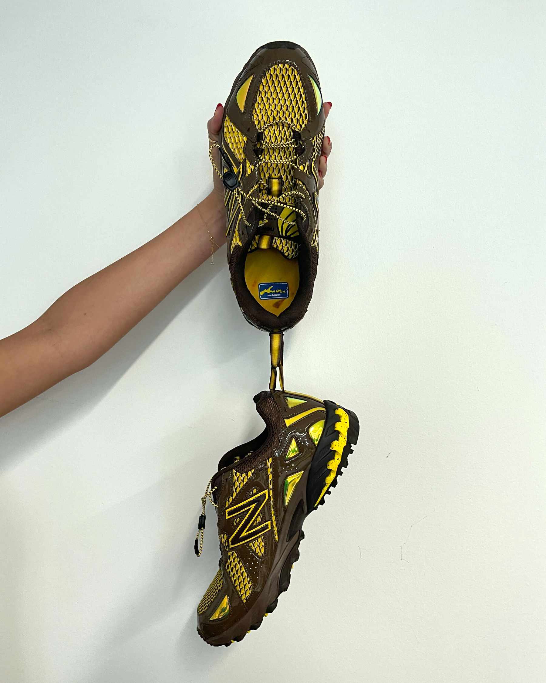 Amine's New Balance 610 shoe, "The Mooz," in brown and yellow colors being held from the rear pull tab