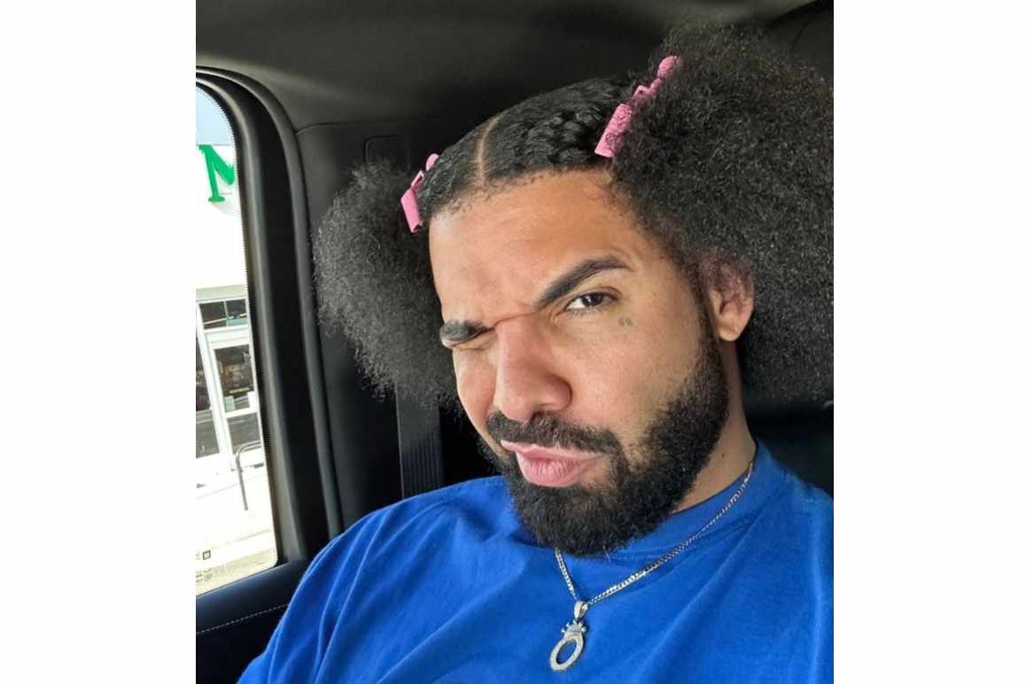 Drake makes a face with his frizzy hair held by pink clips, wearing a blue Fubu T-shirt
