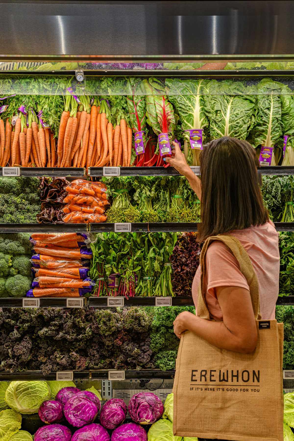 A woman shops at Erewhon Market in California with a tote bag, standing in front of a display of vegetables
