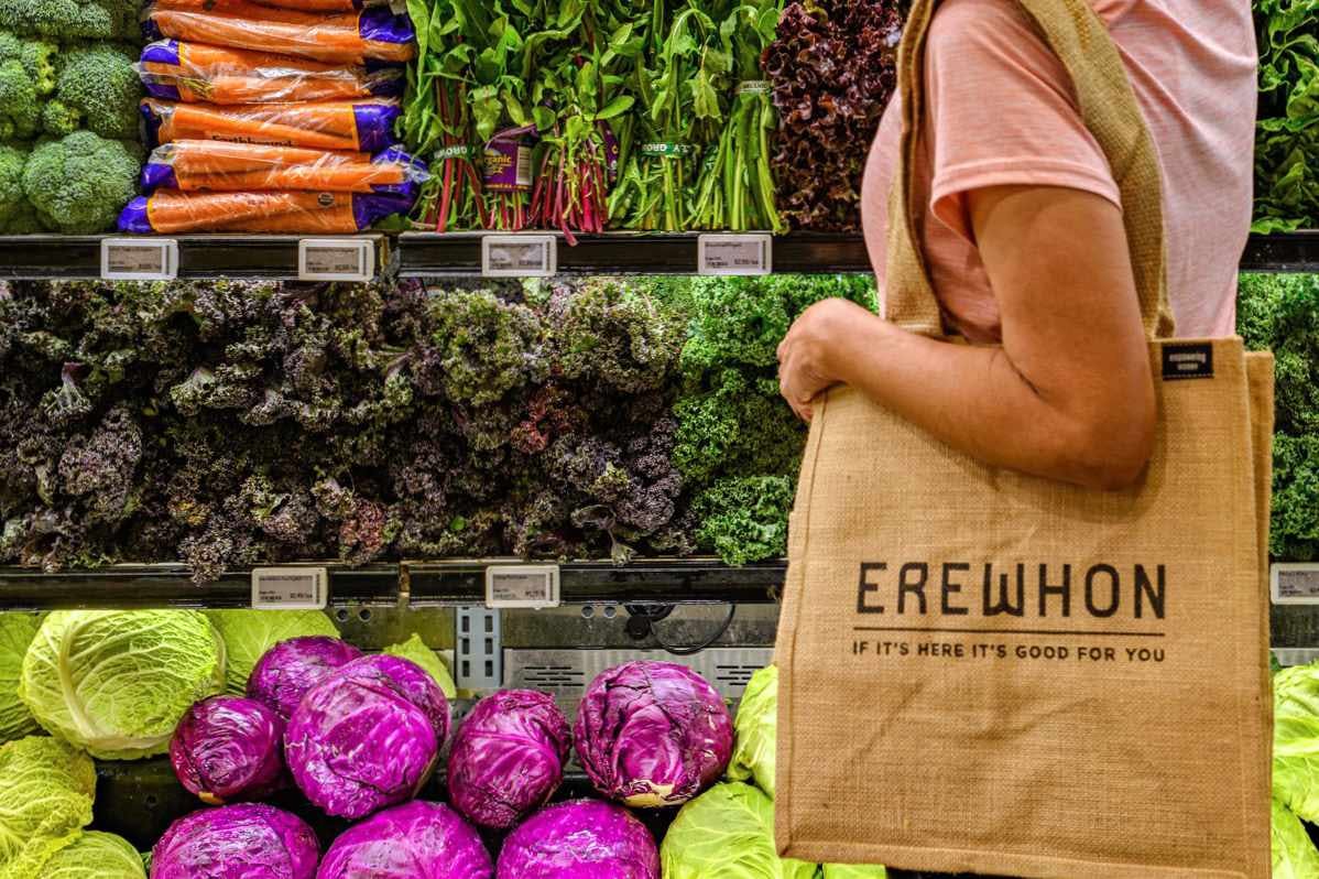 A woman shops at Erewhon Market in California with a tote bag, standing in front of a display of vegetables