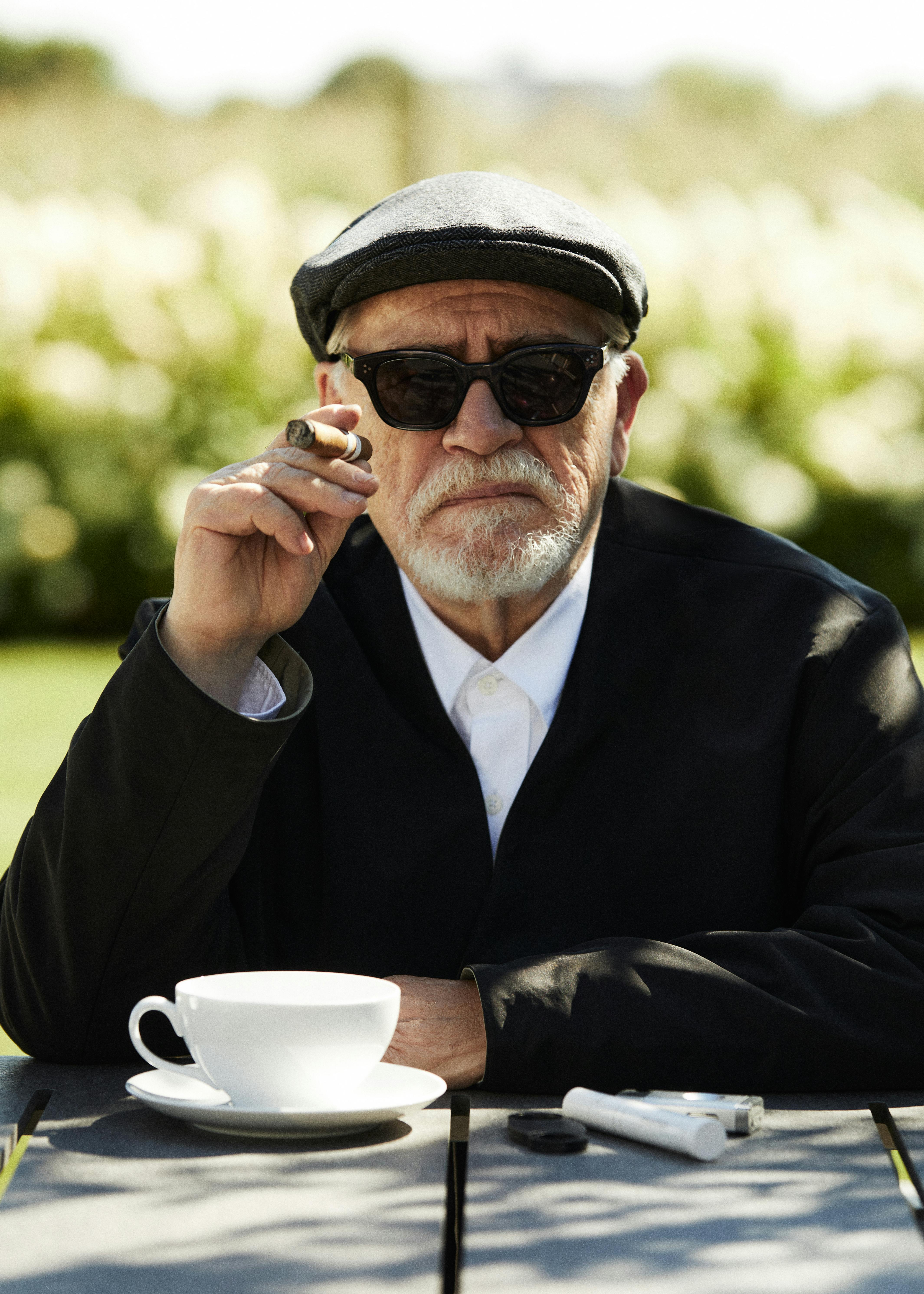 A photograph of Brian Cox, the actor famous for portraying Logan Roy on 'Succession,' wears a black KITH blazer, sunglasses, and white shirt while holding a cigar