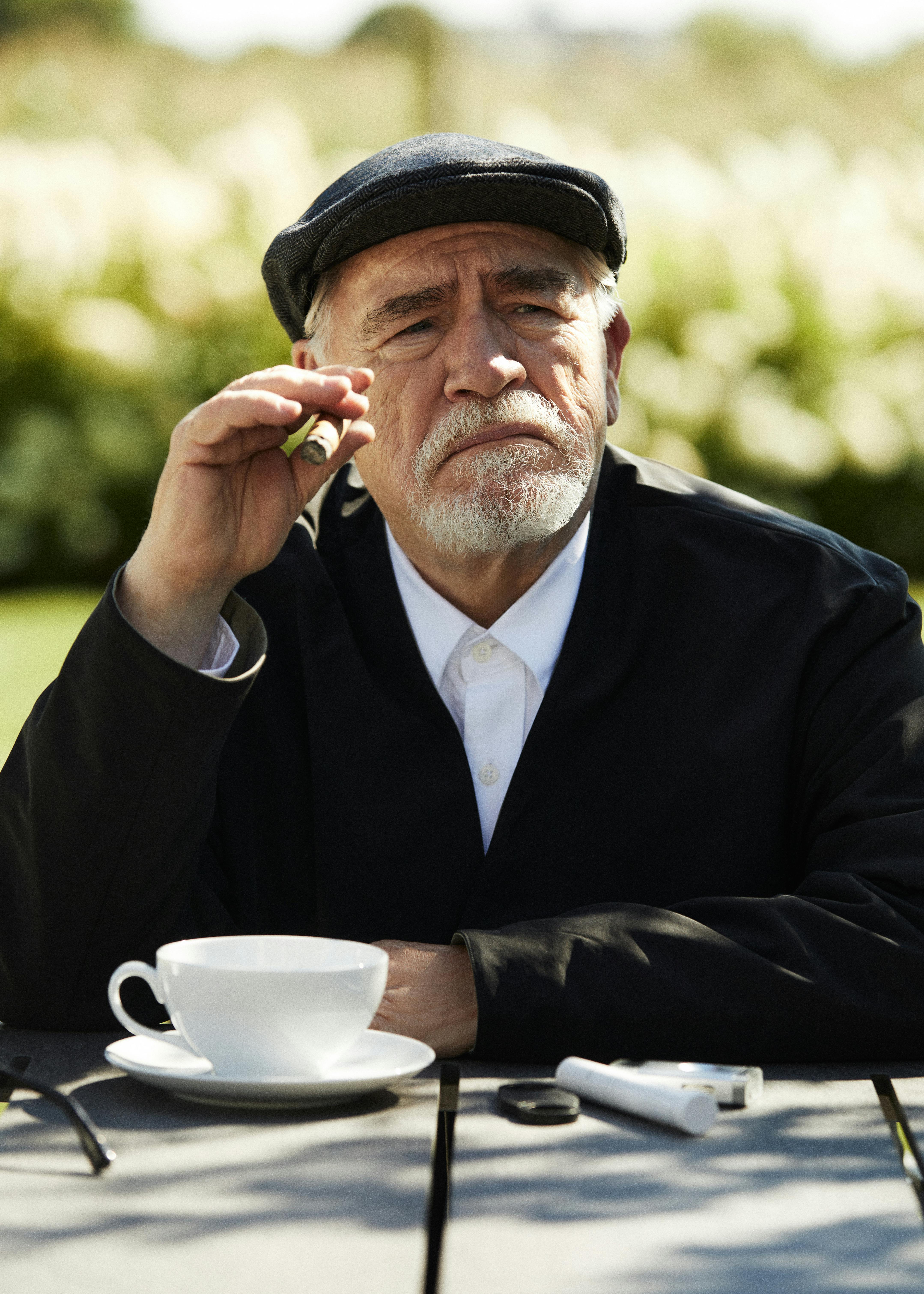 A photograph of Brian Cox, the actor famous for portraying Logan Roy on 'Succession,' wears a black KITH blazer and white shirt while holding a cigar