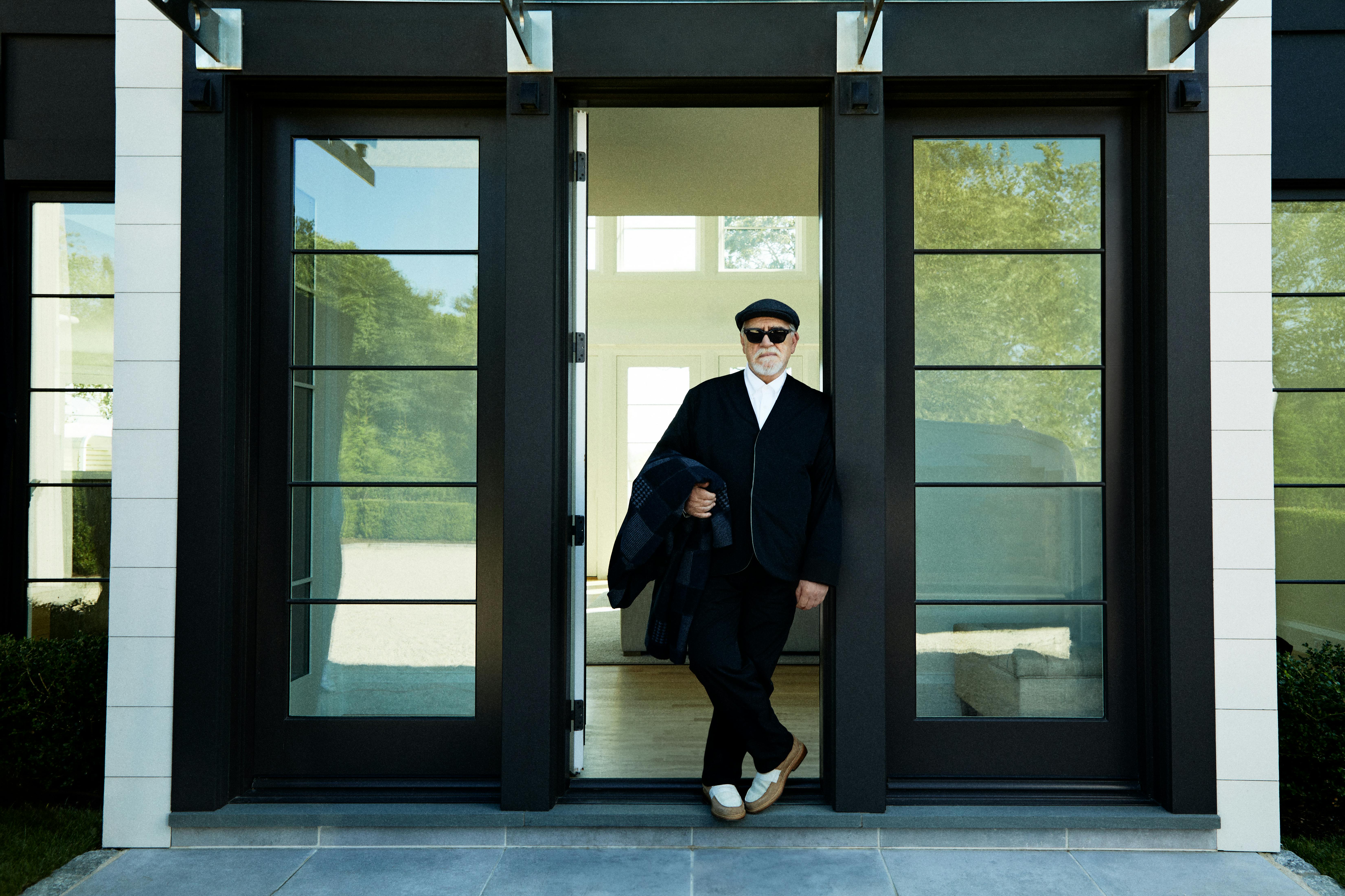 A photograph of Brian Cox, the actor famous for portraying Logan Roy on 'Succession,' wears a black Kith blazer, sunglasses, and white shirt, holding a patchwork coat