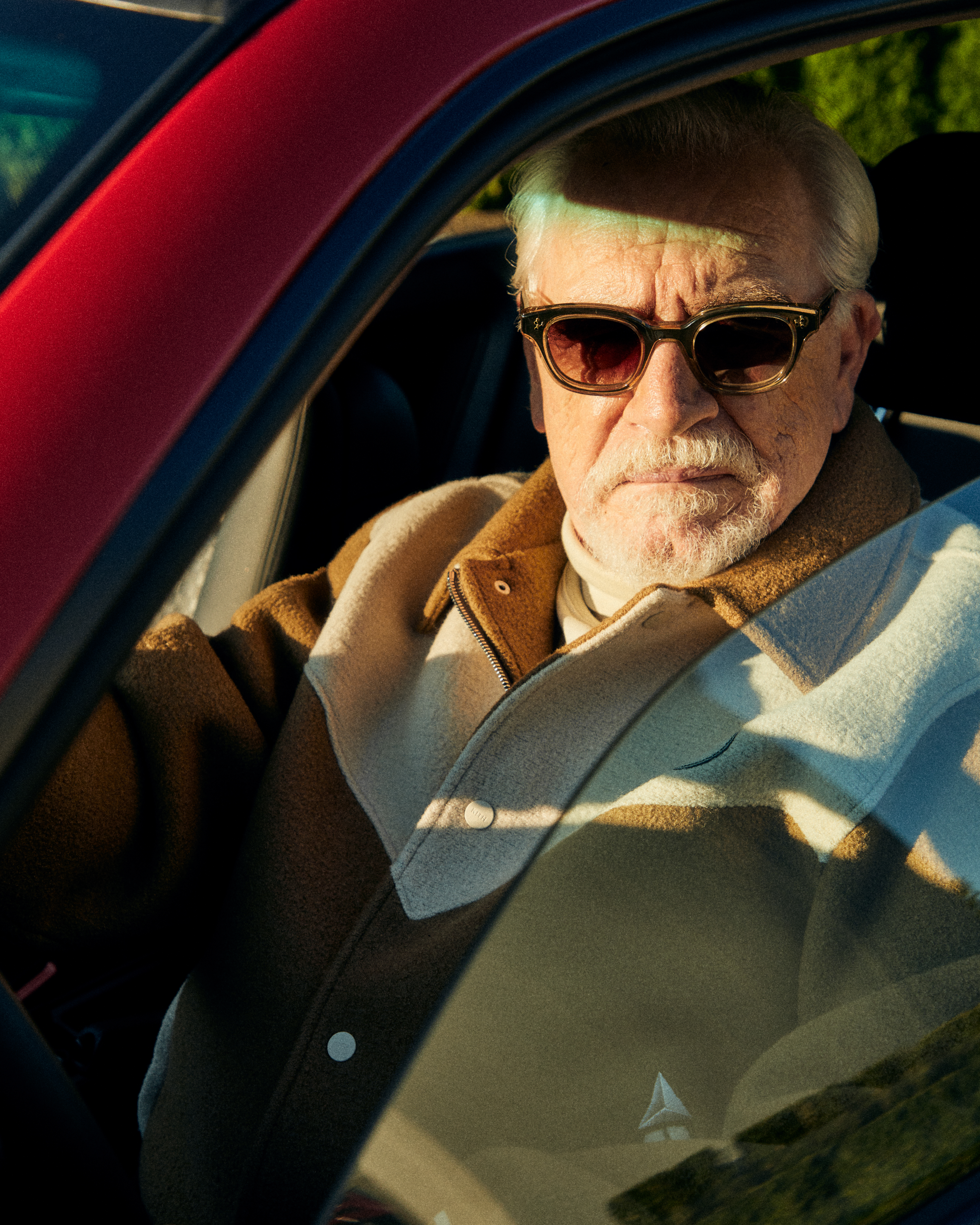 A photograph of Brian Cox, the actor famous for portraying Logan Roy on 'Succession,' wears a beige Kith varsity jacket while sitting in a car