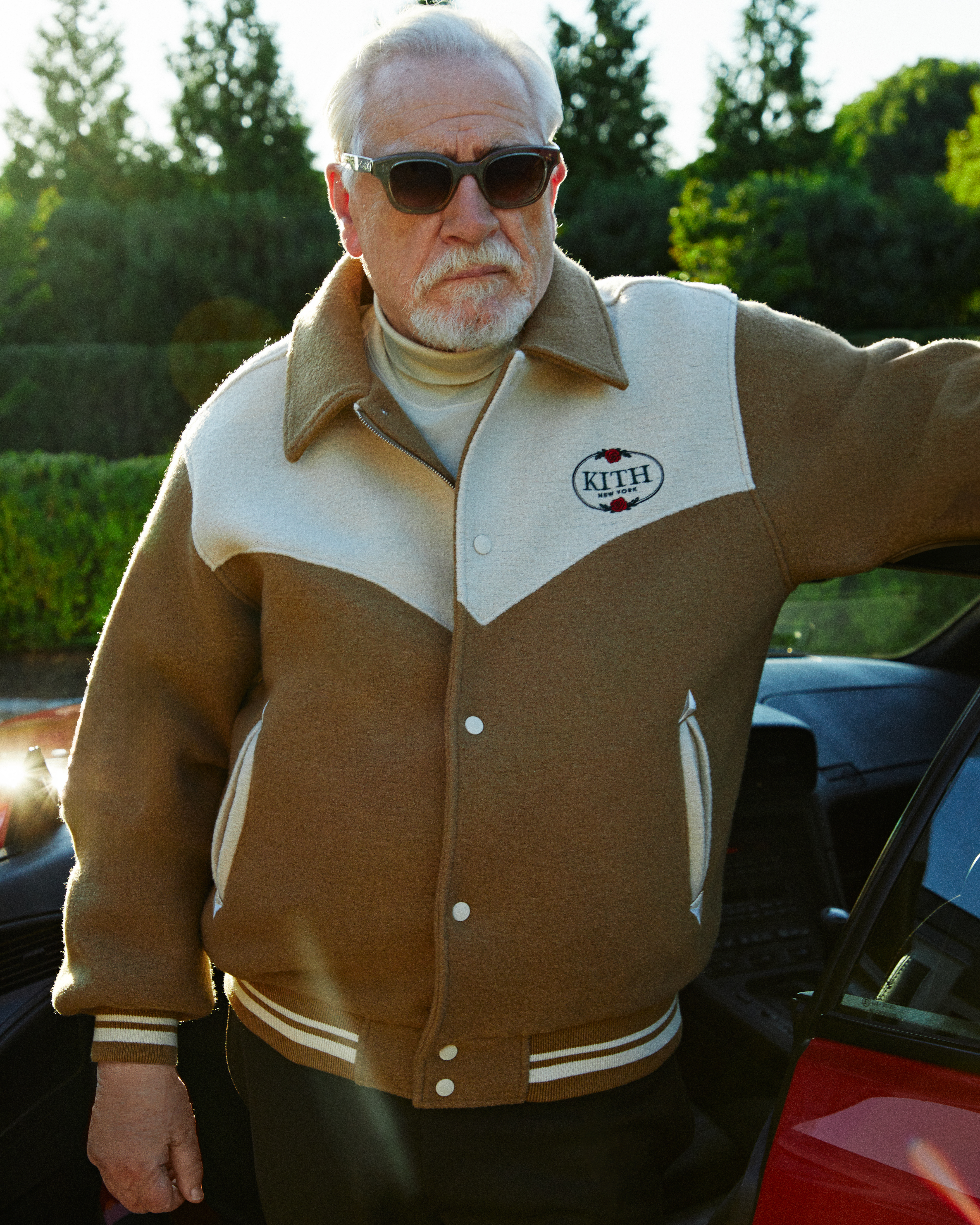 A photograph of Brian Cox, the actor famous for portraying Logan Roy on 'Succession,' wears a beige Kith varsity jacket while standing next to a car