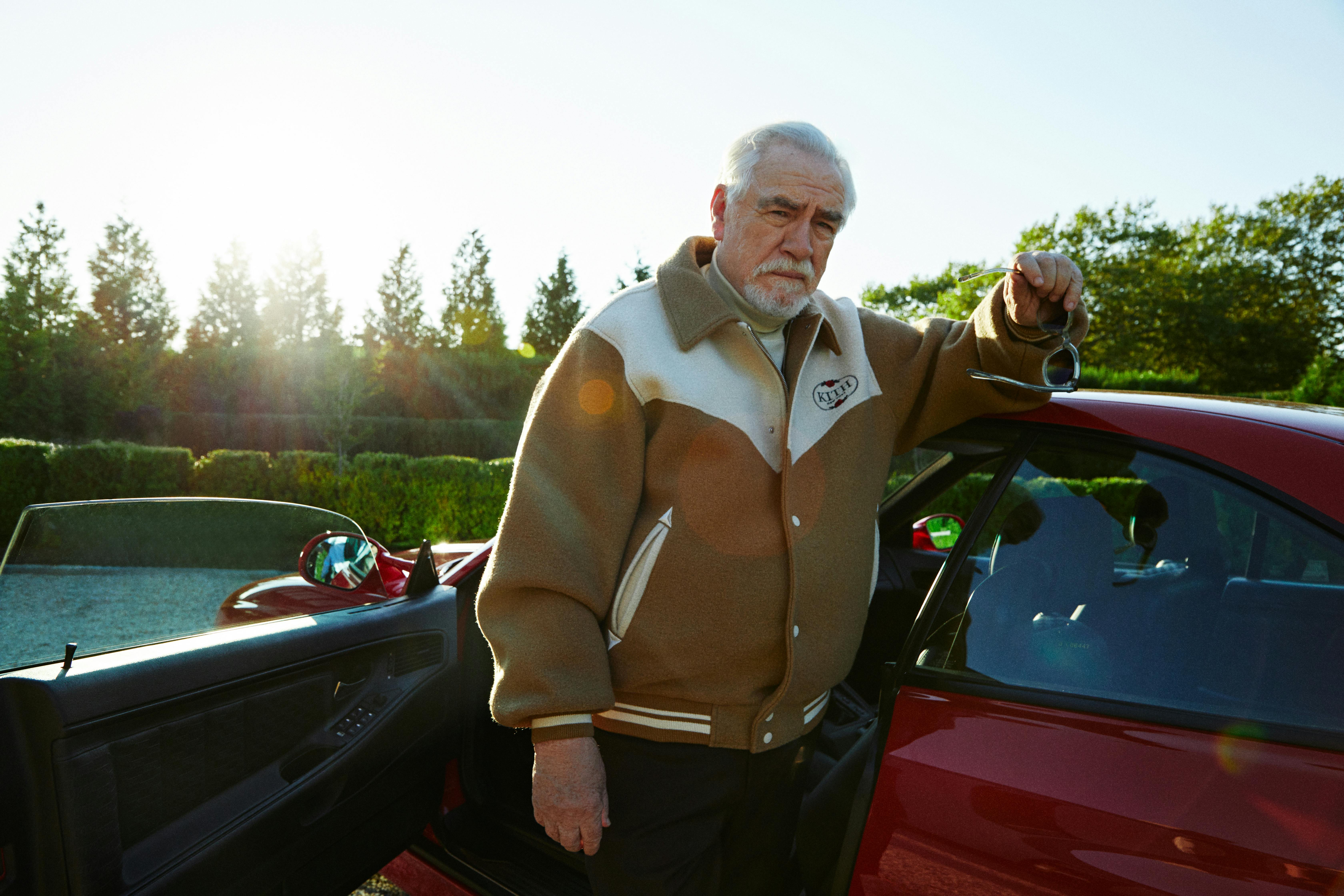 A photograph of Brian Cox, the actor famous for portraying Logan Roy on 'Succession,' wears a beige Kith varsity jacket while standing next to a car