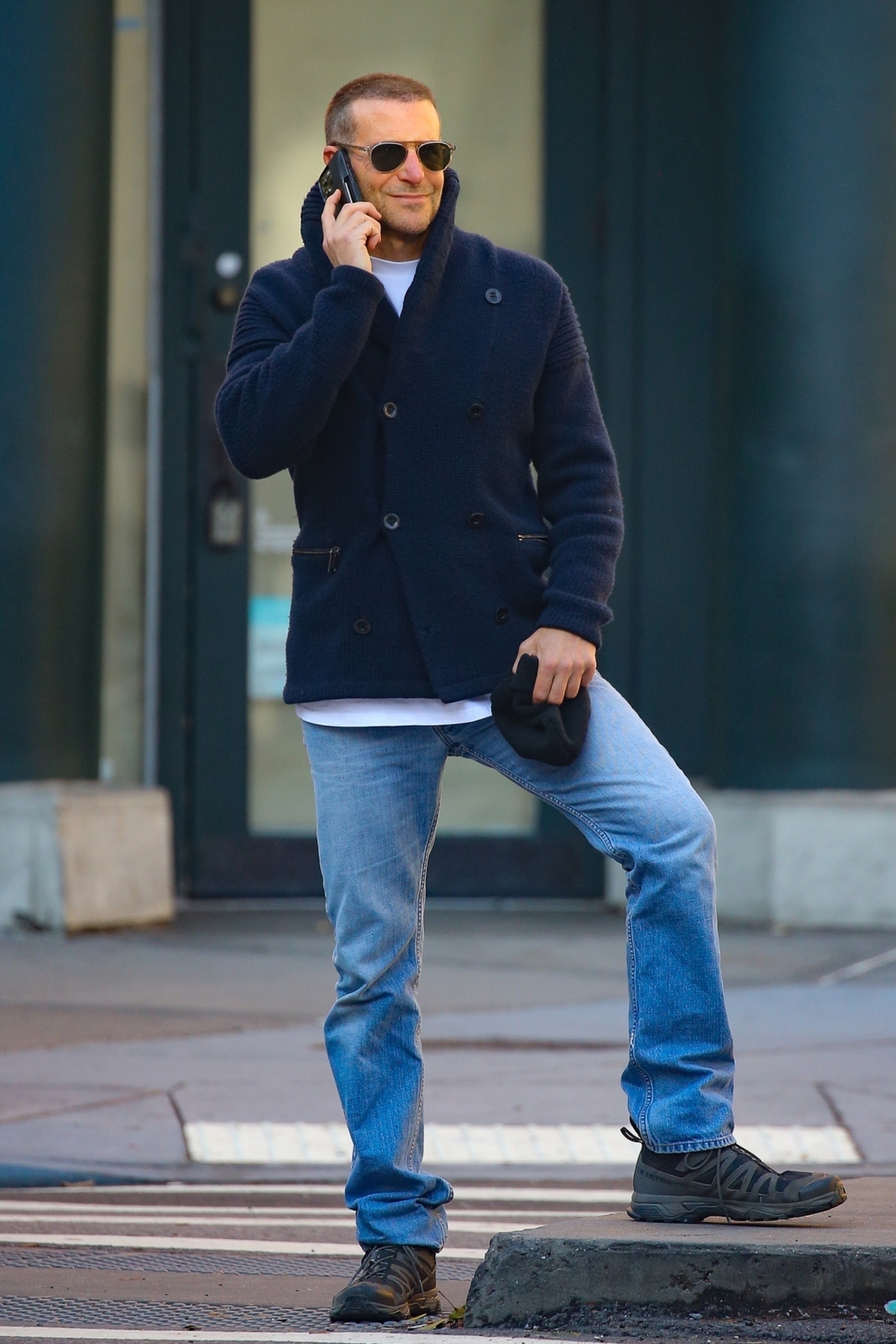Actor Bradley Cooper wears a peacoat, jeans, and Salomon sneakers with sunglasses and a buzzcut hairdo