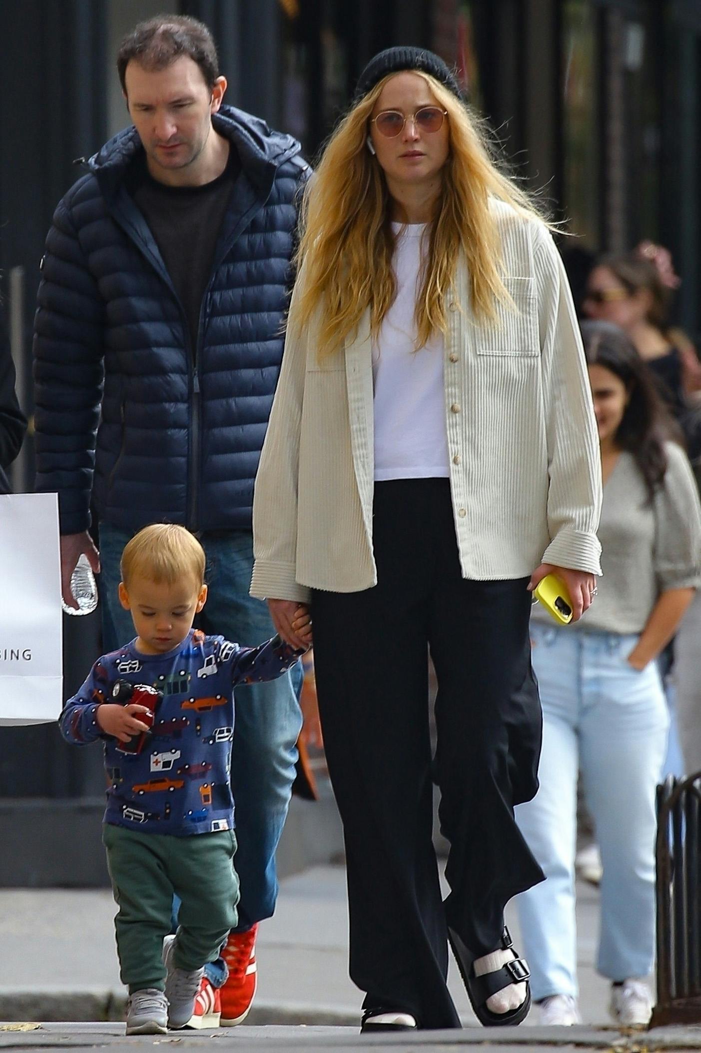 Jennifer Lawrence seen walking in New York with her son wearing a cream corduroy shirt, black slacks, a beanie & socks with sandals