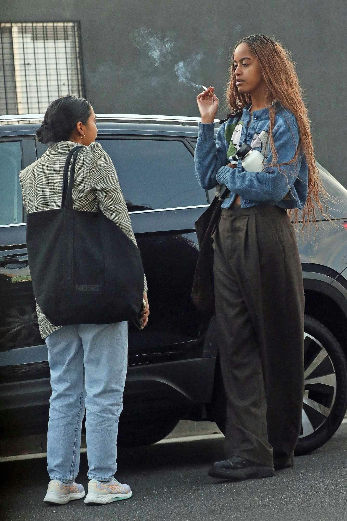 Malia Obama & a friend stand near a car. Obama is smoking, wearing a grey cardigan, brown pants, and black shoes