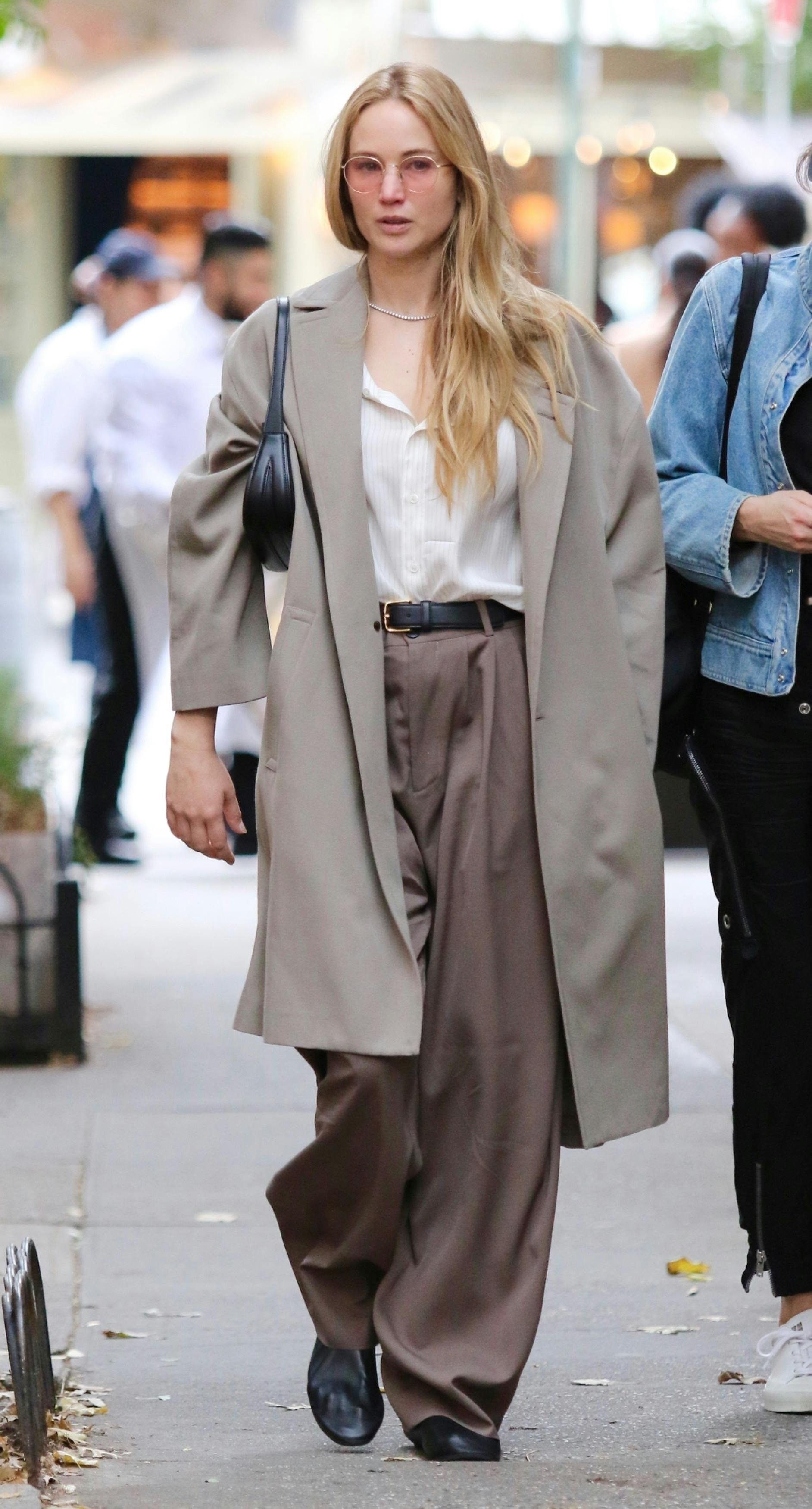 Actress Jennifer Lawrence is seen wearing sunglasses, a beige coat, white shirt, baggy tan pants, and black leather slip-on shoes while walking with Phoebe Waller-Bridge