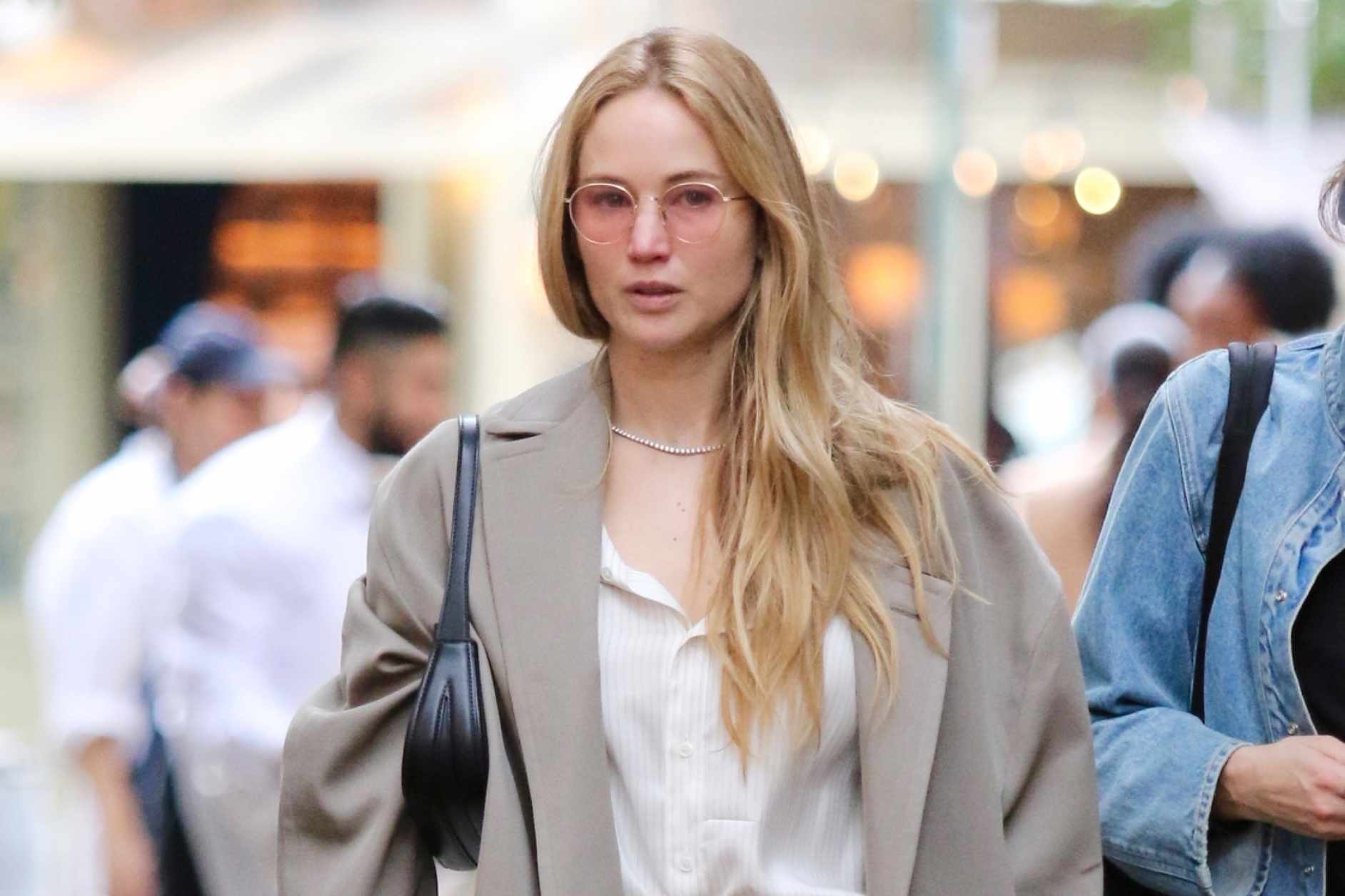 Actress Jennifer Lawrence is seen wearing sunglasses, a beige coat, white shirt, baggy tan pants, and black leather slip-on shoes while walking with Phoebe Waller-Bridge