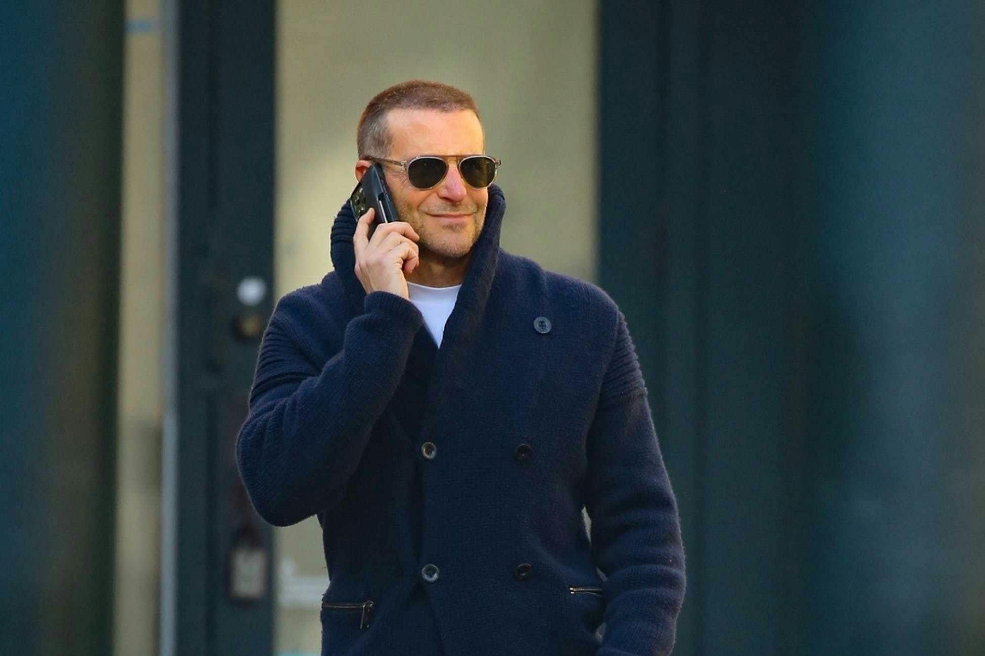 Actor Bradley Cooper wears a peacoat, jeans, and Salomon sneakers with sunglasses and a buzzcut hairdo