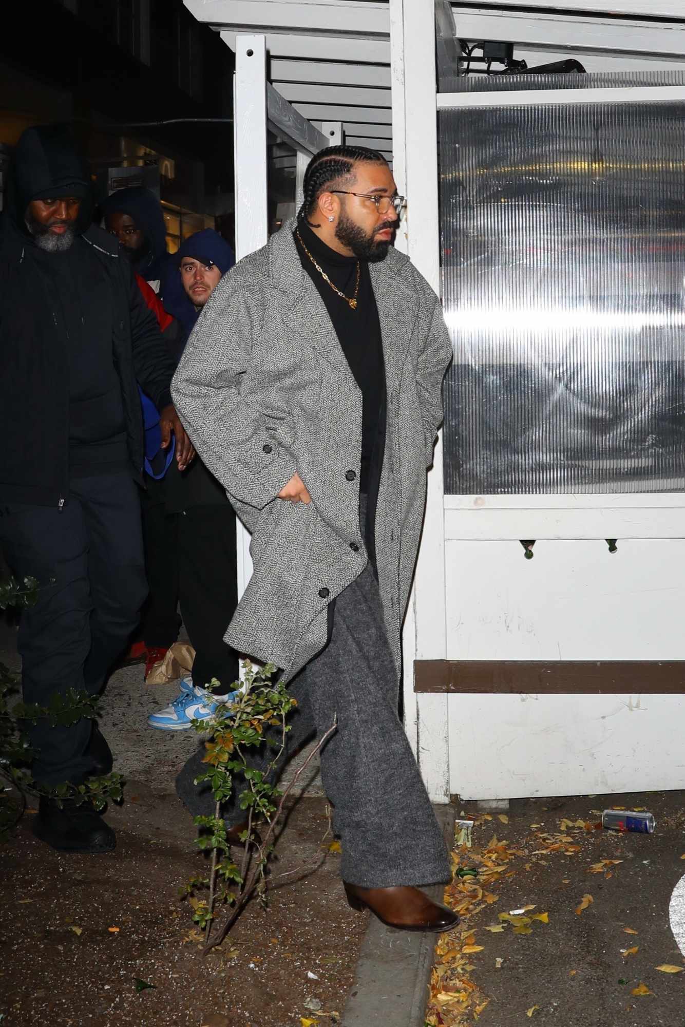 Drake wears a braids, glasses, a grey coat, wide pants & brown boots