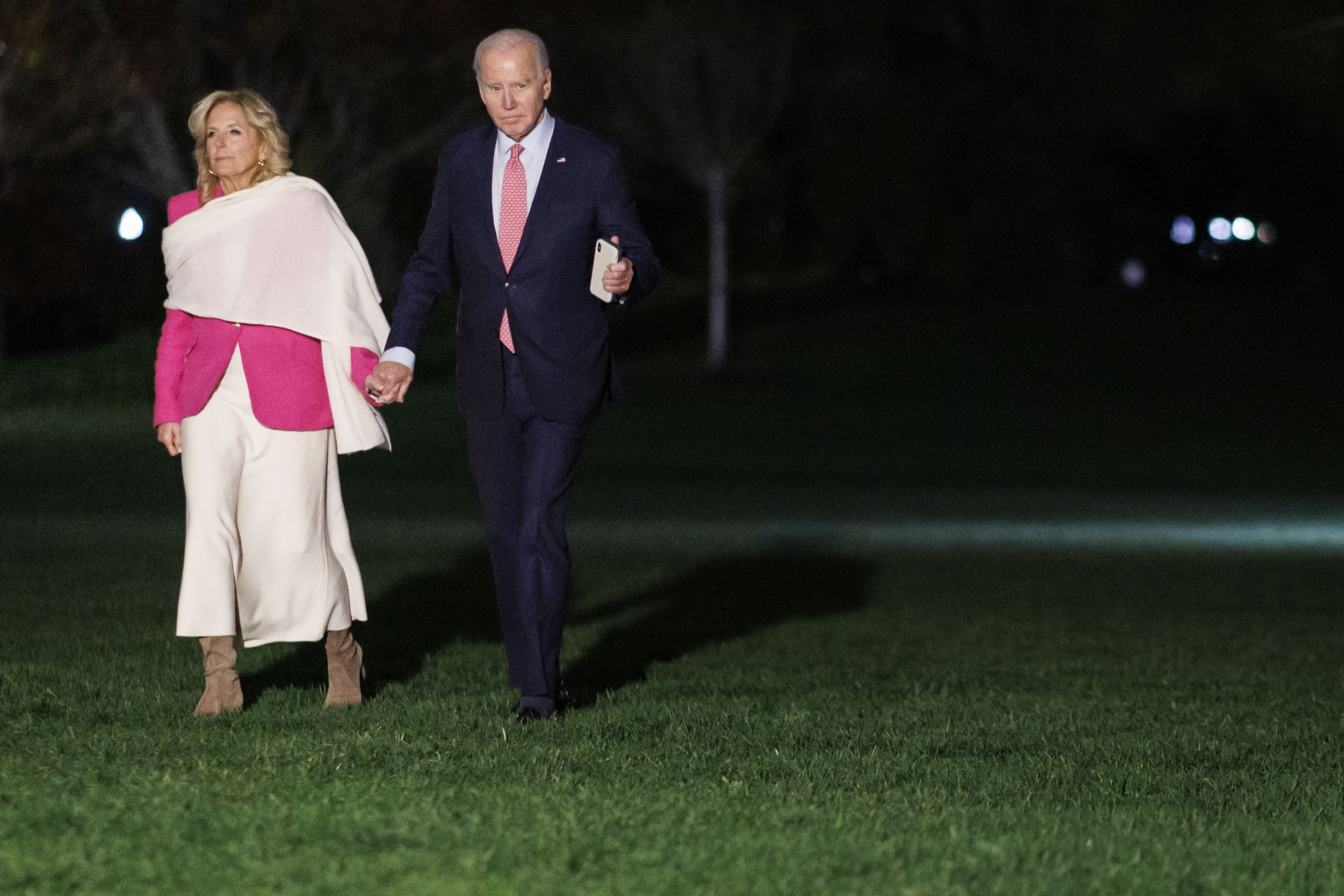 Jill Biden wears a giant white scarf over a pink blazer while holding hands with Joe Biden
