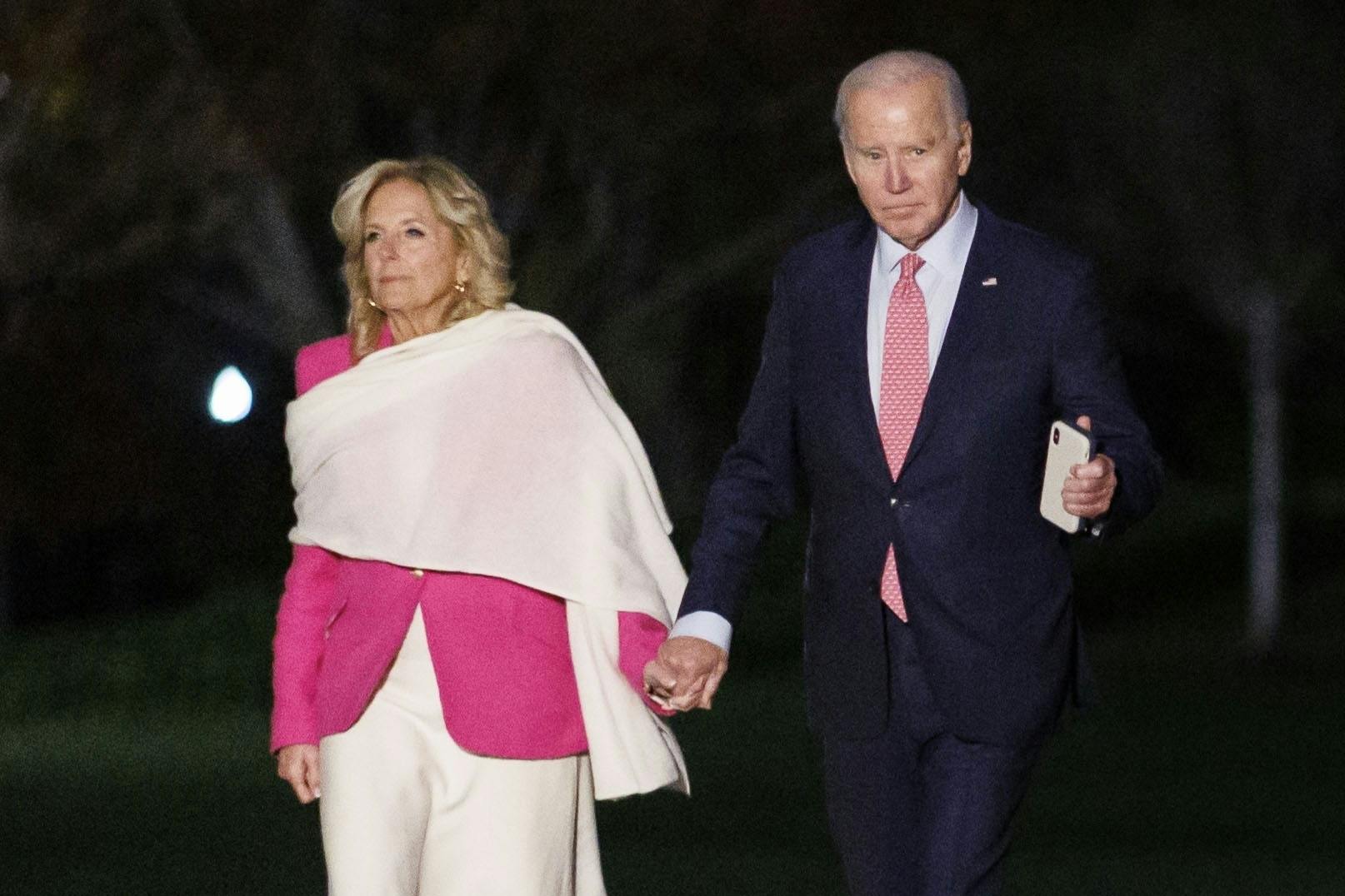 Jill Biden wears a giant white scarf over a pink blazer while holding hands with Joe Biden