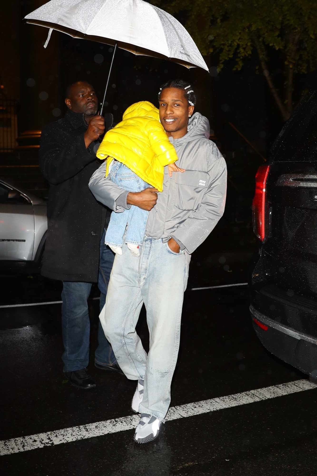 A$AP Rocky smiles while wearing a grey Miu Miu jacket & jeans with son RZA wearing a yellow puffer jacket & Onitsuka Tiger sneakers