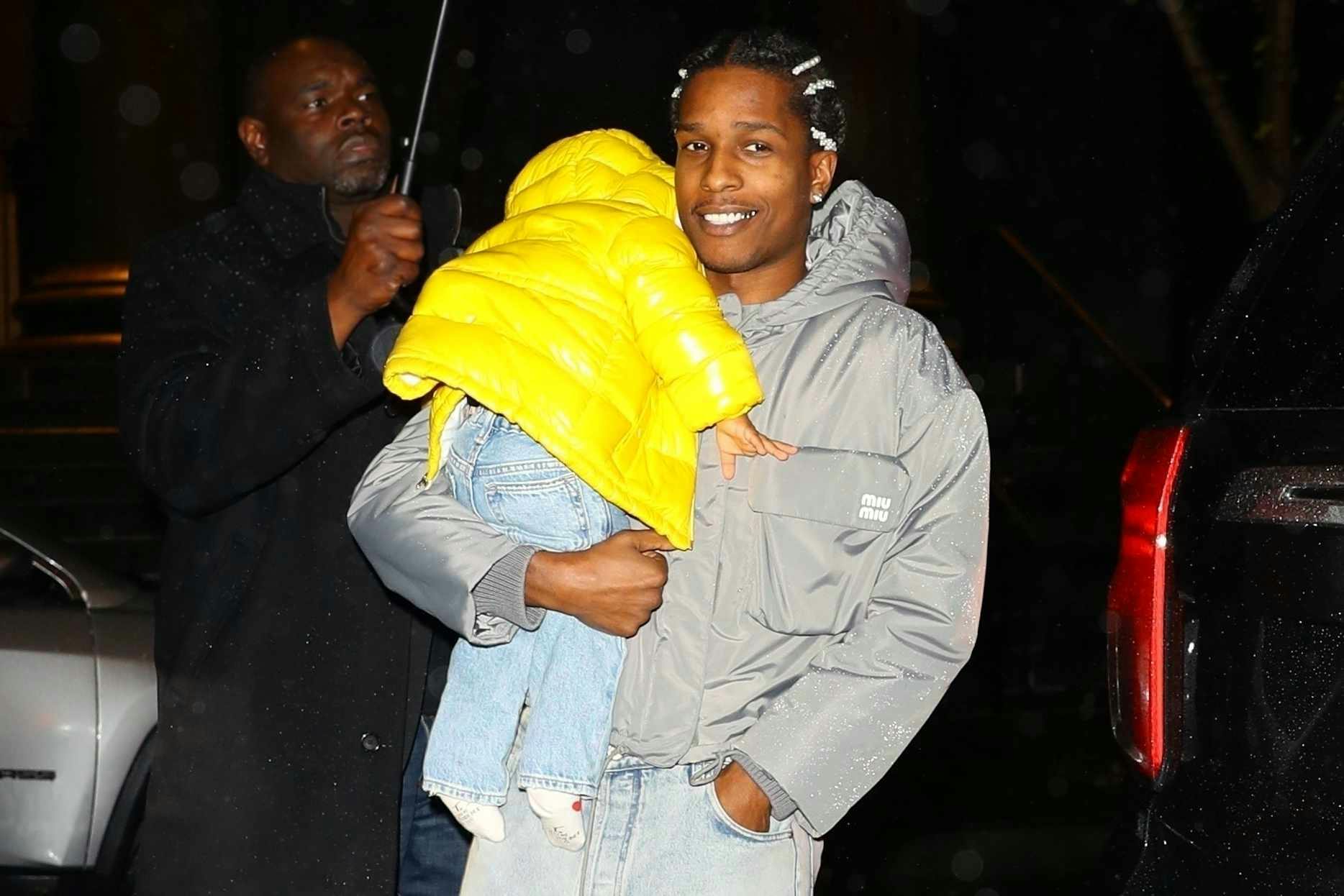 A$AP Rocky smiles while wearing a grey Miu Miu jacket & jeans with son RZA wearing a yellow puffer jacket & Onitsuka Tiger sneakers