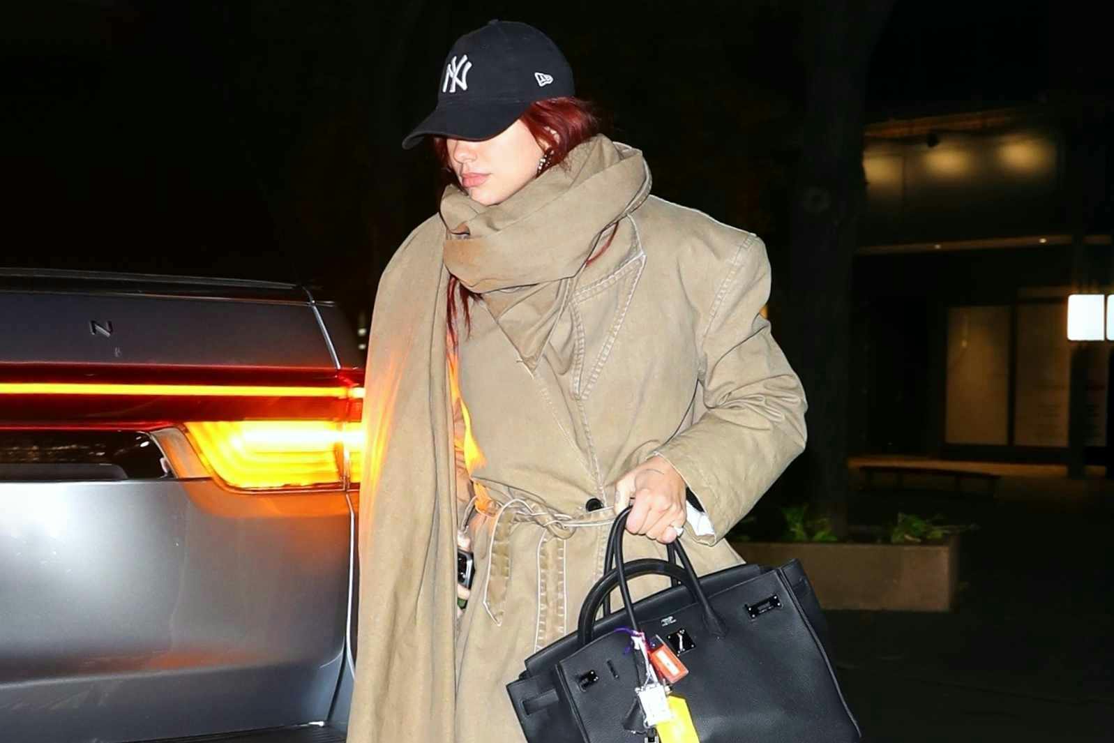 Dua Lipa wears a beige scarf, coat, Yankees hat, PUMA Palermo sneakers, and black Hermes bag