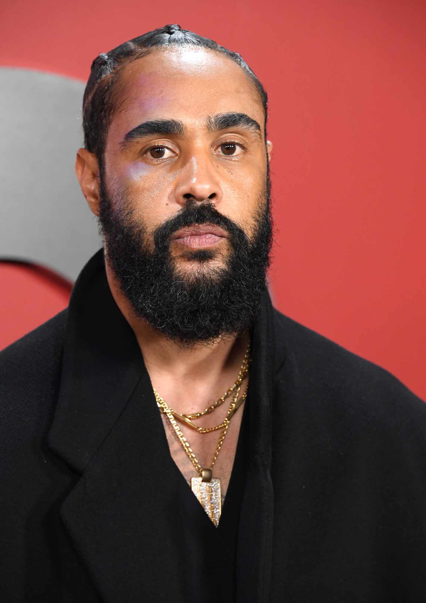 Fear of God Jerry Lorenzo wears a black coat and gold jewelry in front of a red backdrop