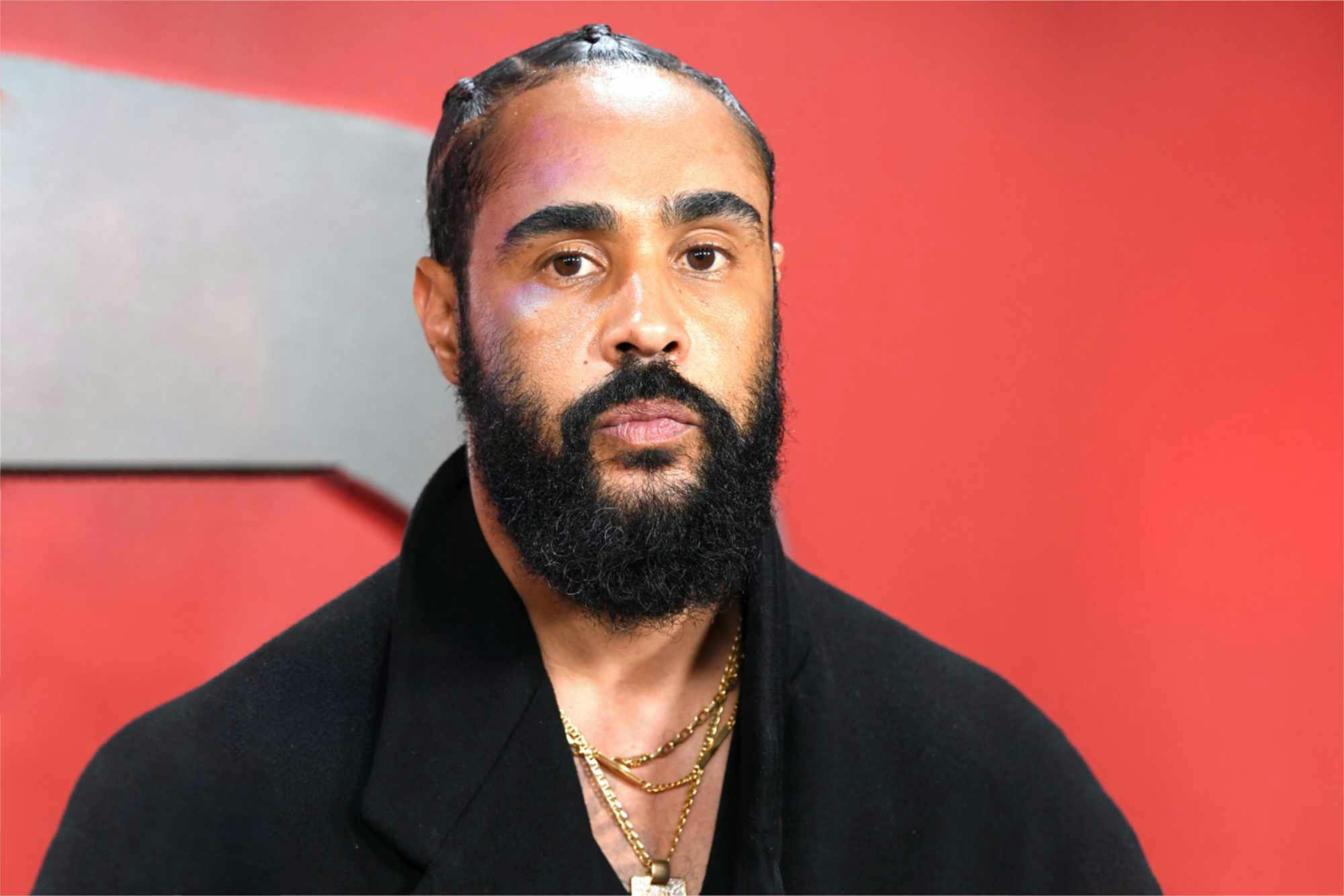 Fear of God Jerry Lorenzo wears a black coat and gold jewelry in front of a red backdrop