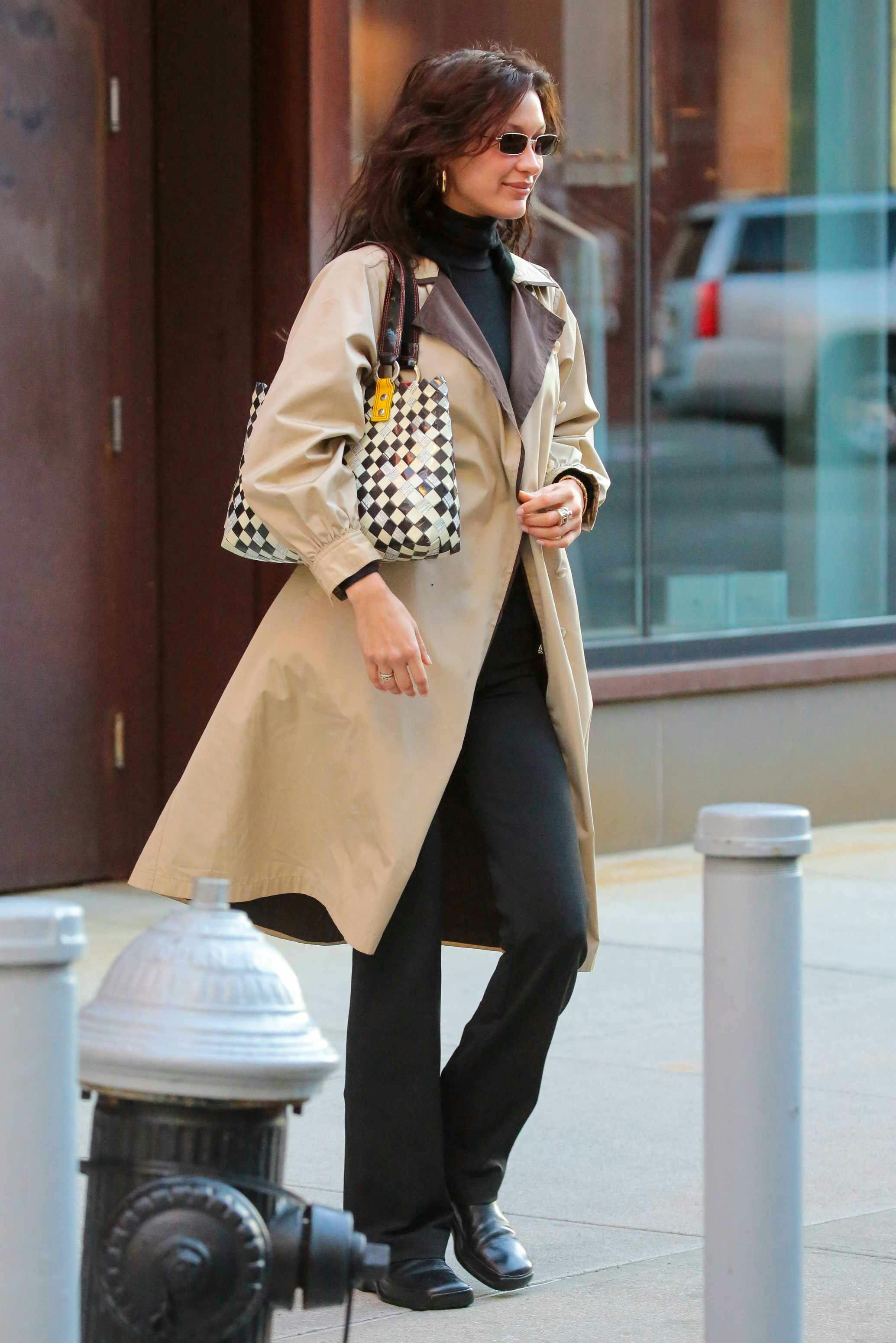 Bella Hadid wears a beige trench coat, black pants, small sunglasses, and a gold sequin bag