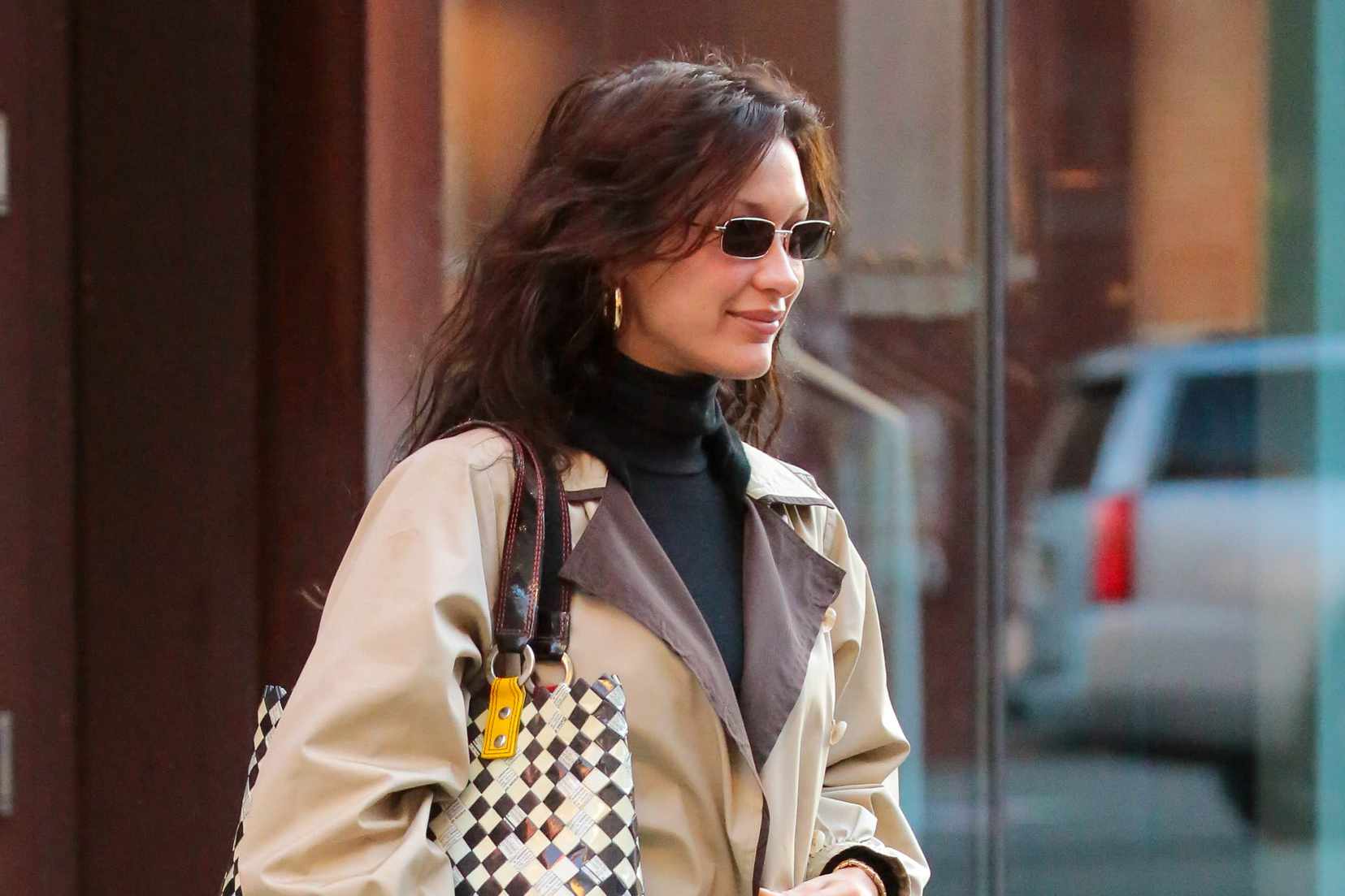 Bella Hadid wears a beige trench coat, black pants, small sunglasses, and a gold sequin bag