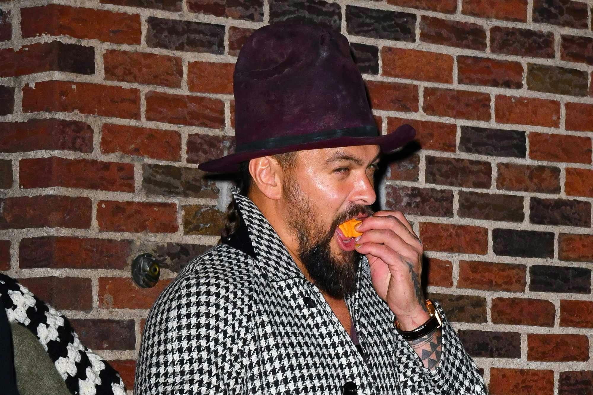 Jason Momoa wears a tall hat, houndstooth coat & slippers while eating Cheez-Its at The Late Show studios