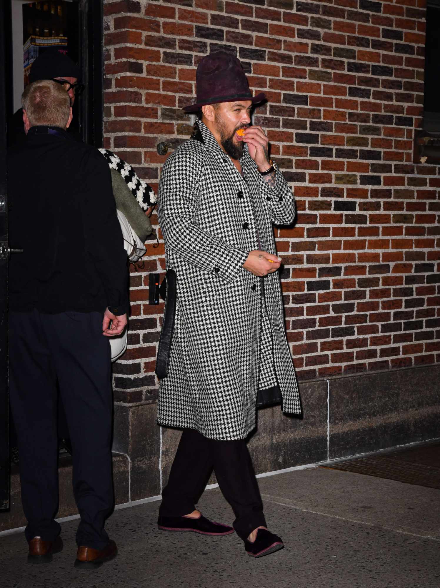 Jason Momoa wears a tall hat, houndstooth coat & slippers while eating Cheez-Its at The Late Show studios