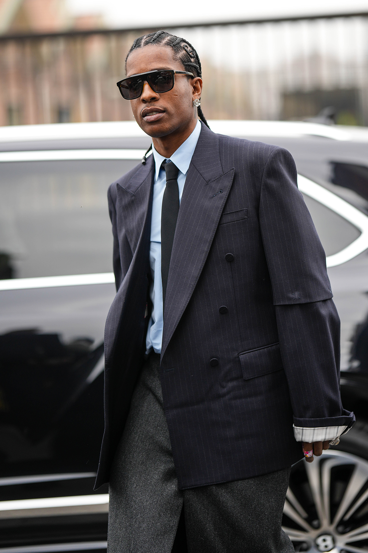 Asap Rocky wears black sunglasses, a white shirt, a black tie, a black striped print pattern oversized blazer jacket, a gray slit / split long skirt, matching gray suit pants, rings, outside Gucci, during the Milan Fashion Week Womenswear Fall/Winter 2023/2024 on February 24, 2023 in Milan, Italy.