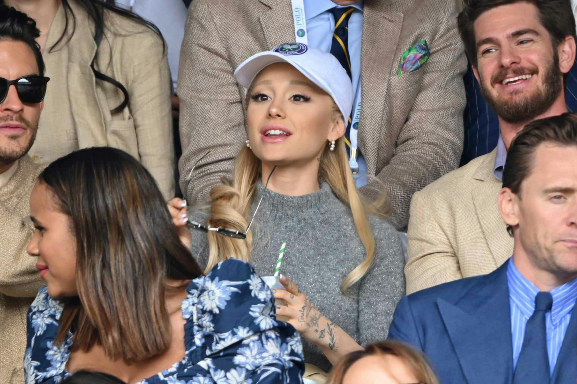 Ariana Grande wears a white cap over blonde hair and grey sweater at the US Open