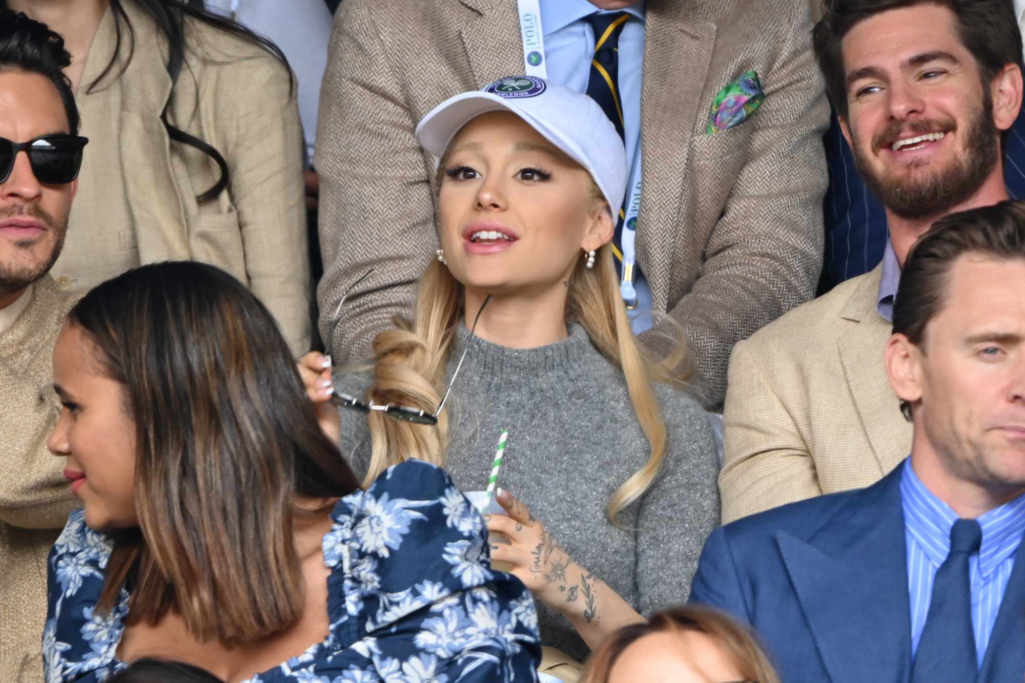 Ariana Grande wears a white cap over blonde hair and grey sweater at the US Open