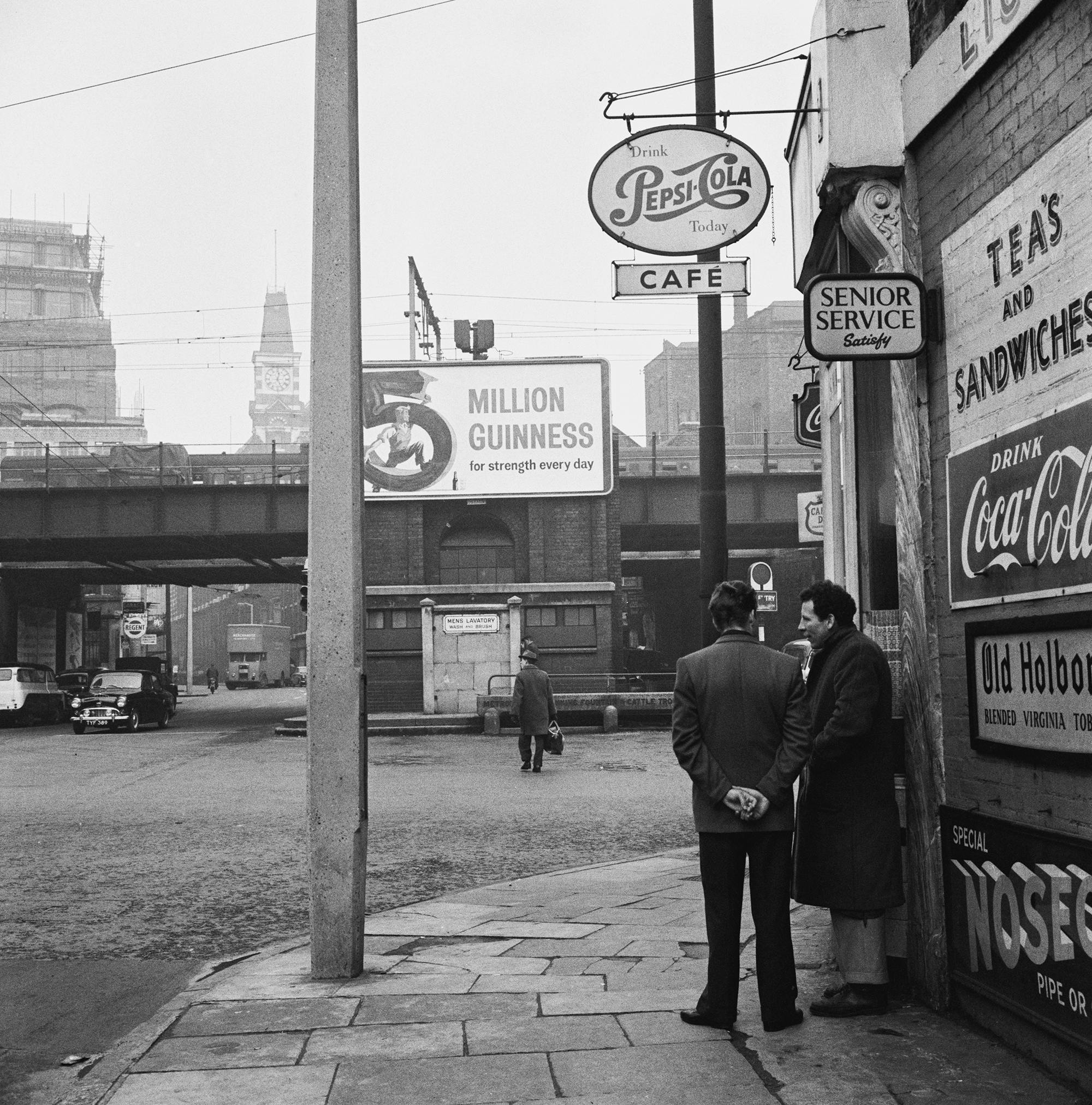 Two men stand on a street corner near advertising billboards around Brick Lane in East London, 1960.