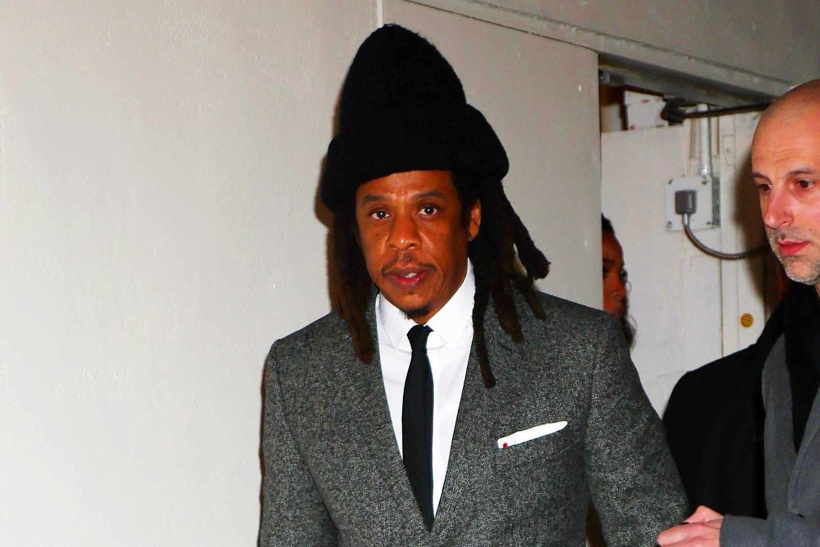 JAY-Z wears a tall black beanie and grey suit with white shirt at the mea culpa movie premiere