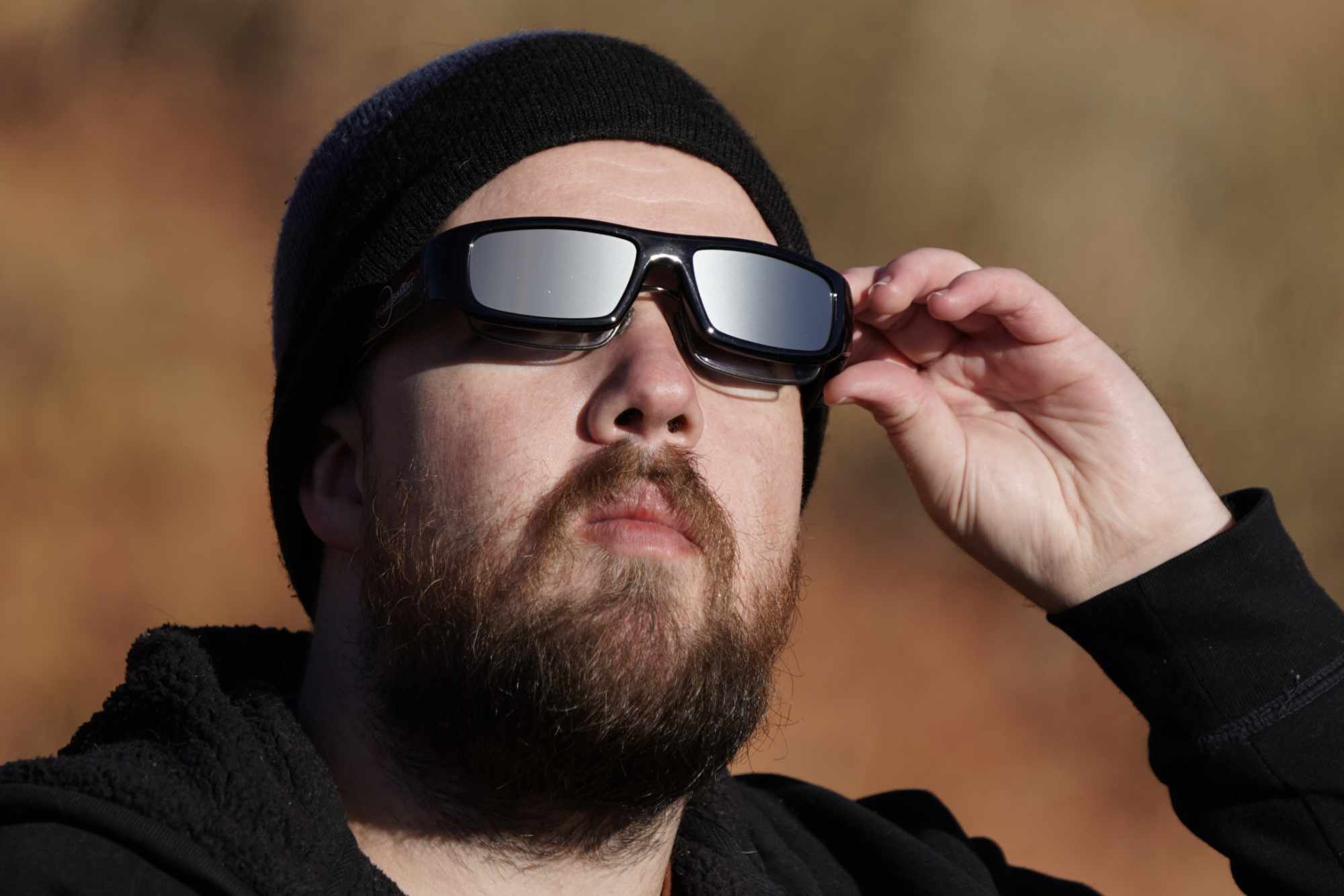 Black and silver eclipse glasses worn by a man with a beard and dark beanie hat