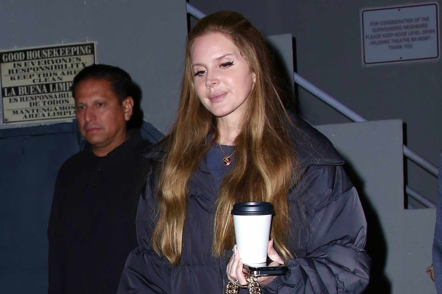 Lana Del Rey wears a black puffer jacket, leggings, and white keds with a black leather chanel bag