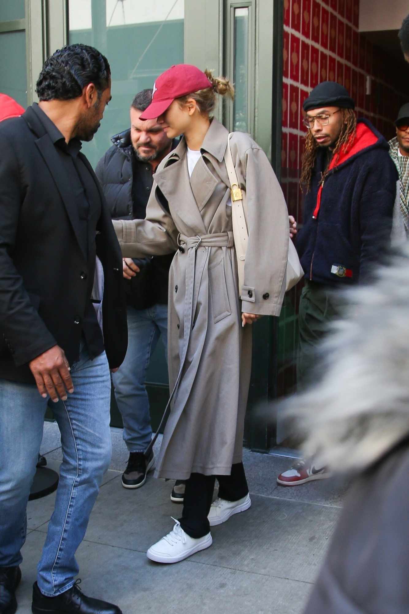 Zendaya wears a white Louis Vuitton bag, red hat, beige trench coat, black leggings, and white On Roger Federer sneakers