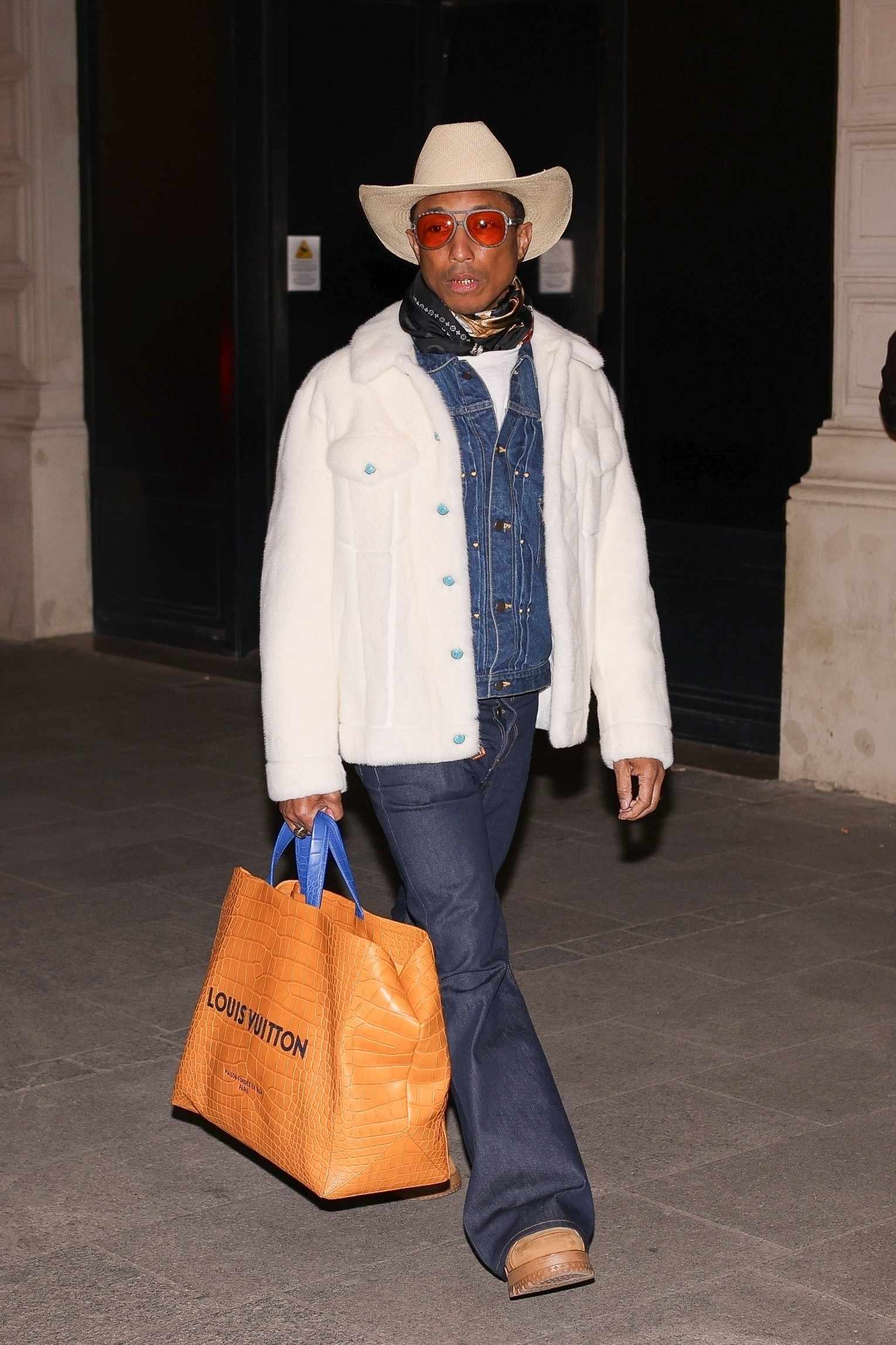 Pharrell Williams wears a white cowboy hat, white shearling jacket, Louis Vuitton leather bag, and denim western shirt with bootcut jeans