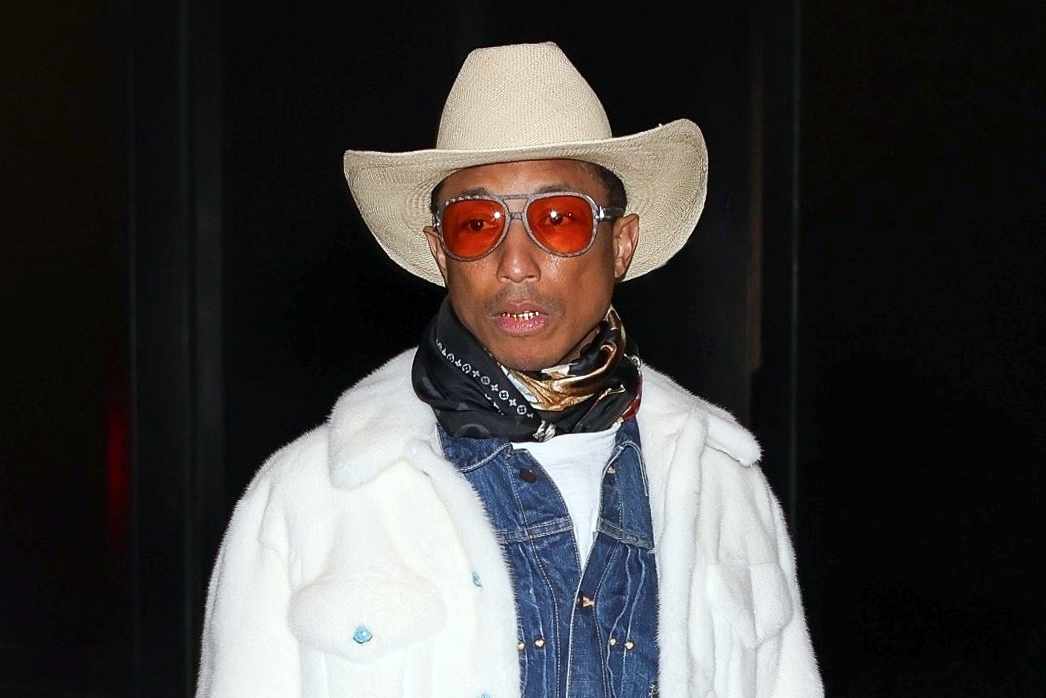 Pharrell Williams wears a white cowboy hat, white shearling jacket, Louis Vuitton leather bag, and denim western shirt with bootcut jeans