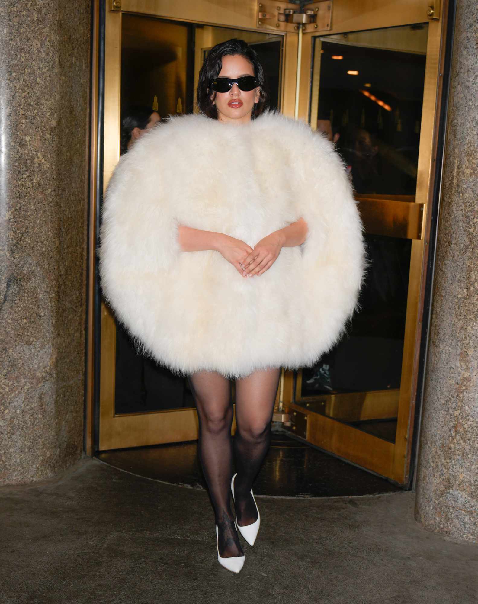Rosalia wears a furry white Celine coat with black sunglasses, tights, and white heels