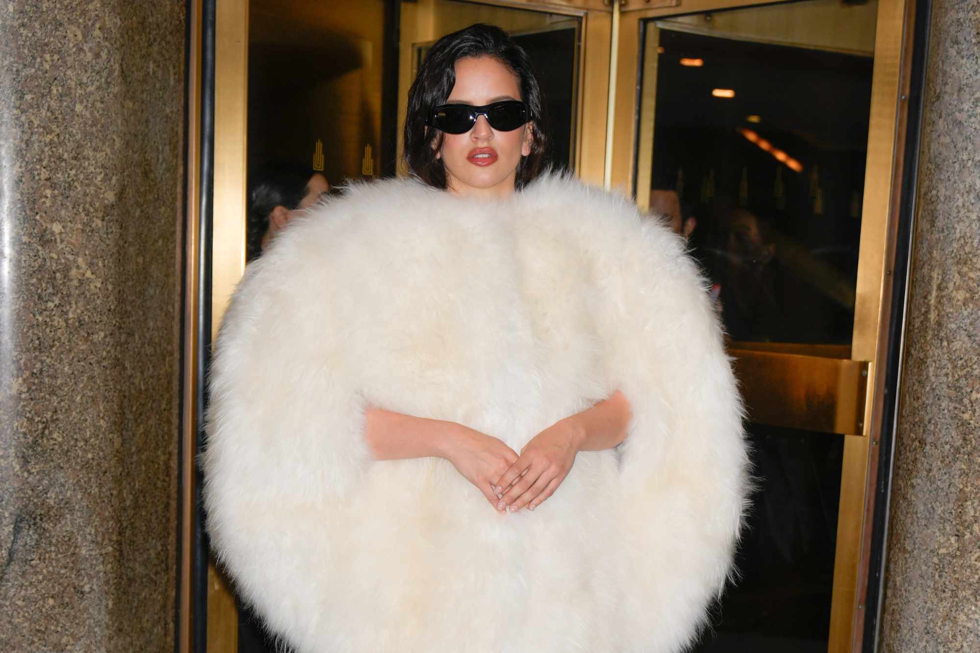 Rosalia wears a furry white Celine coat with black sunglasses, tights, and white heels