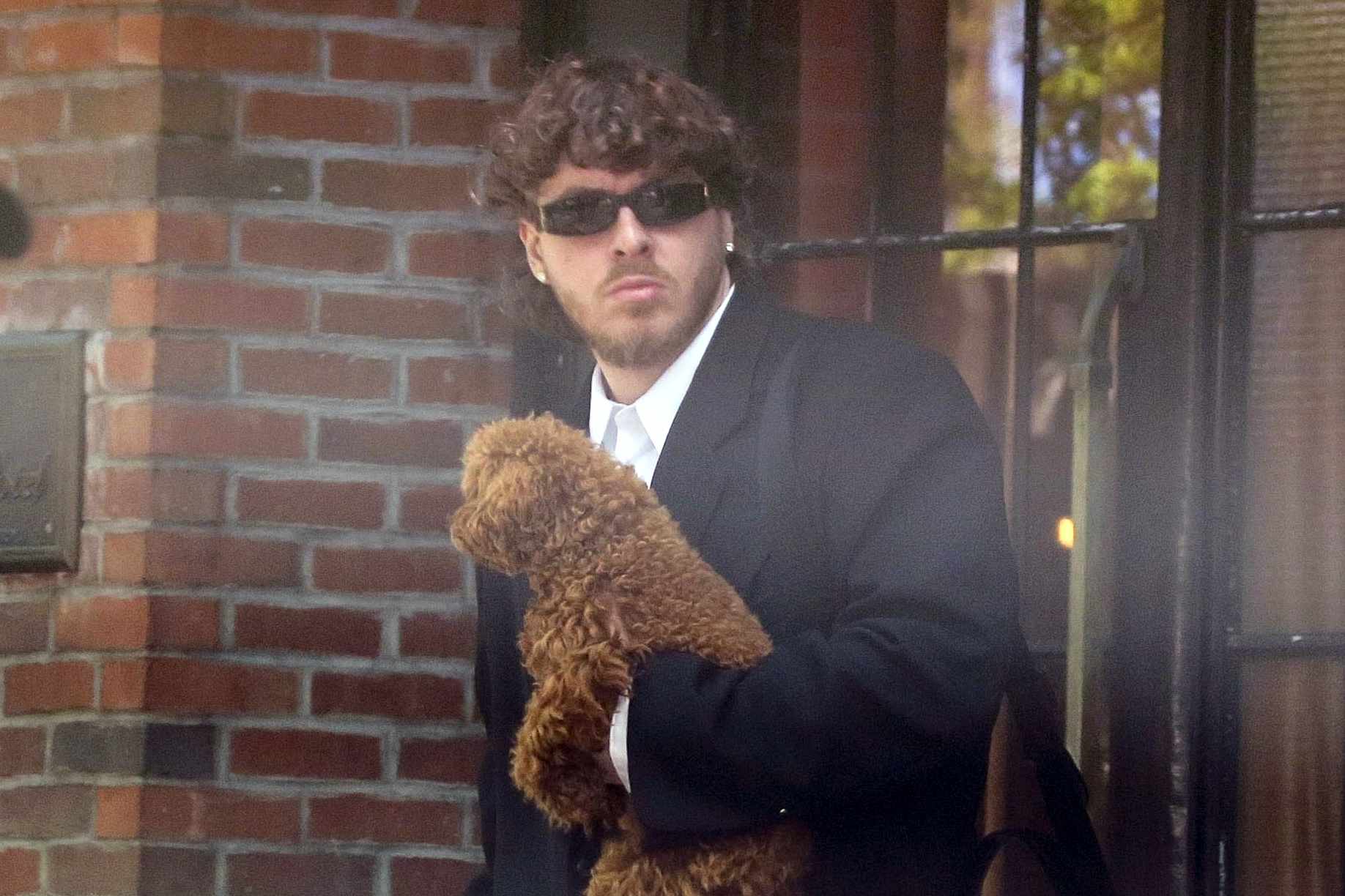 Jack Harlow wears dark sunglasses, a black suit, and his curly-haired dog