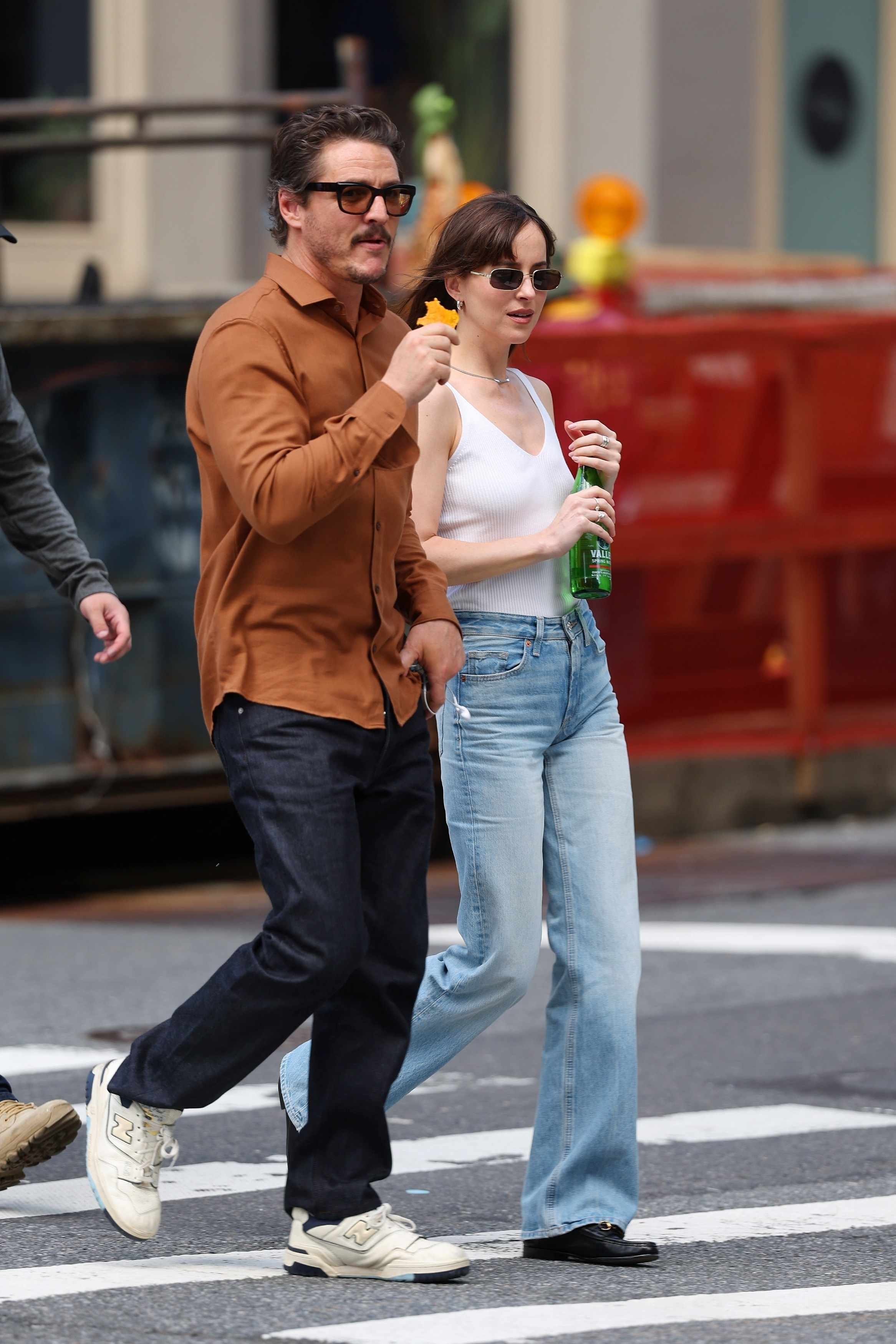 Pedro Pascal wears sunglasses, a red shirt, black pants and white ALD New Balance sneakers with Dakota Johnson, who wears black sunglasses, a white top, blue jeans, and loafers