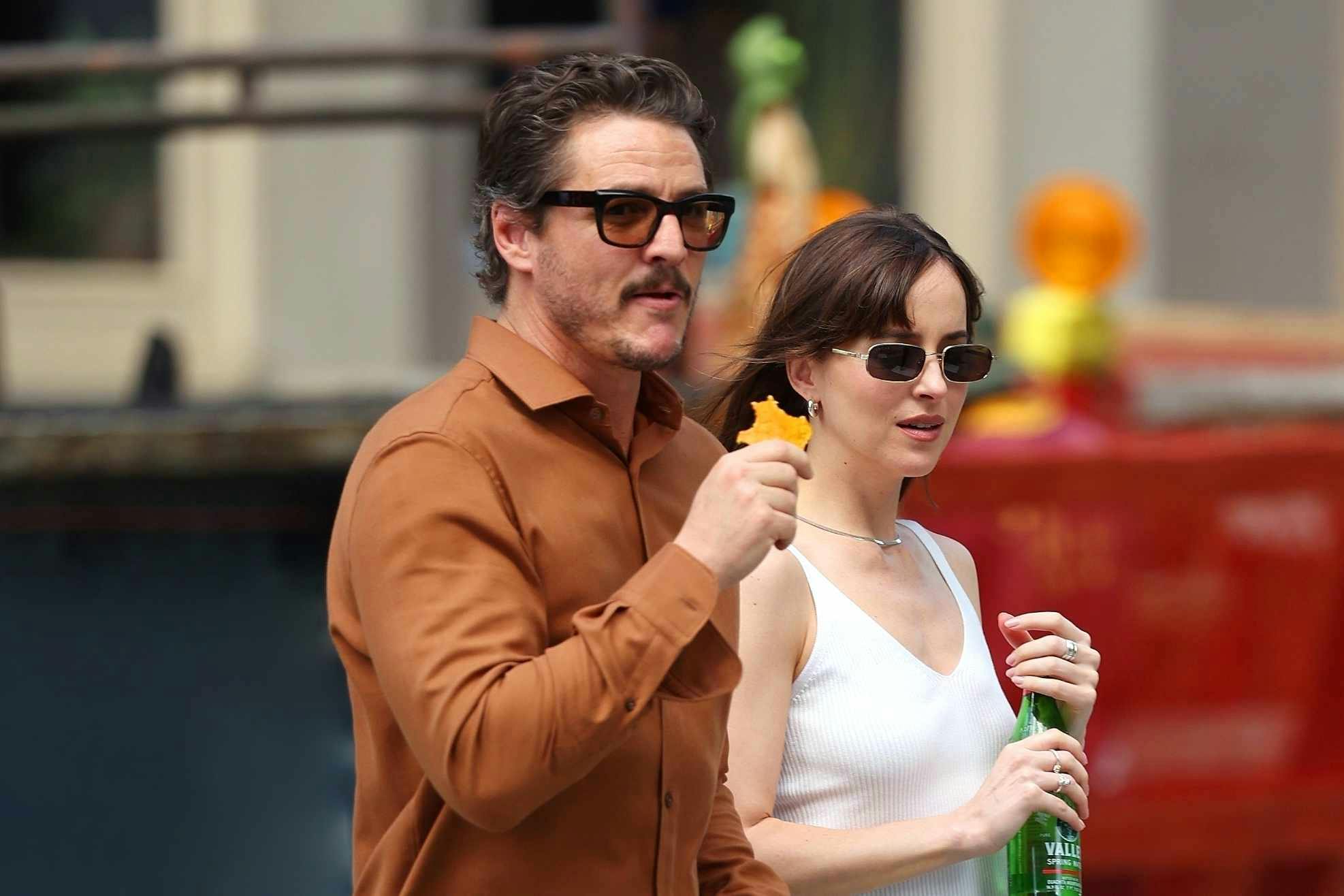 Pedro Pascal wears sunglasses, a red shirt, black pants and white ALD New Balance sneakers with Dakota Johnson, who wears black sunglasses, a white top, blue jeans, and loafers