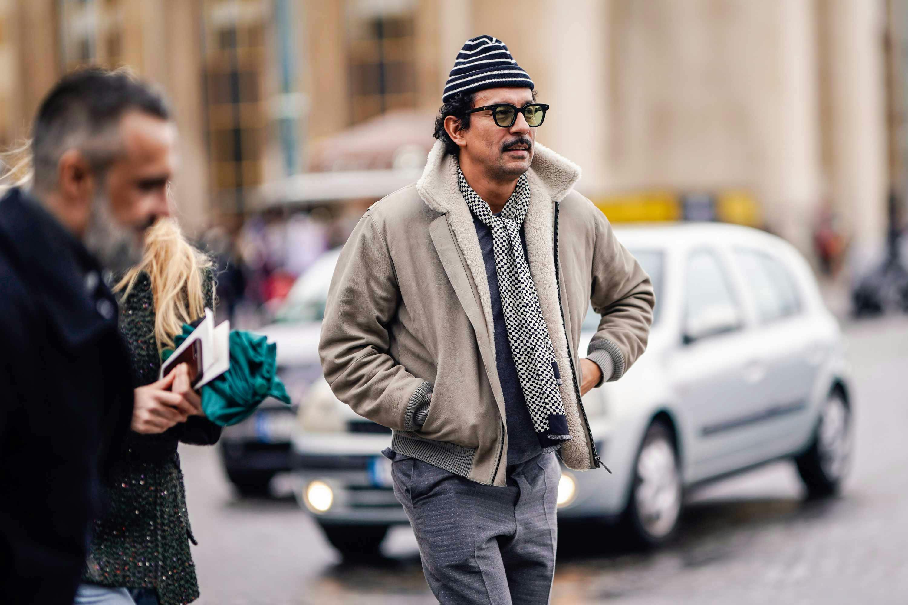 designer Haider Ackermann wears a grey jacket and striped hat