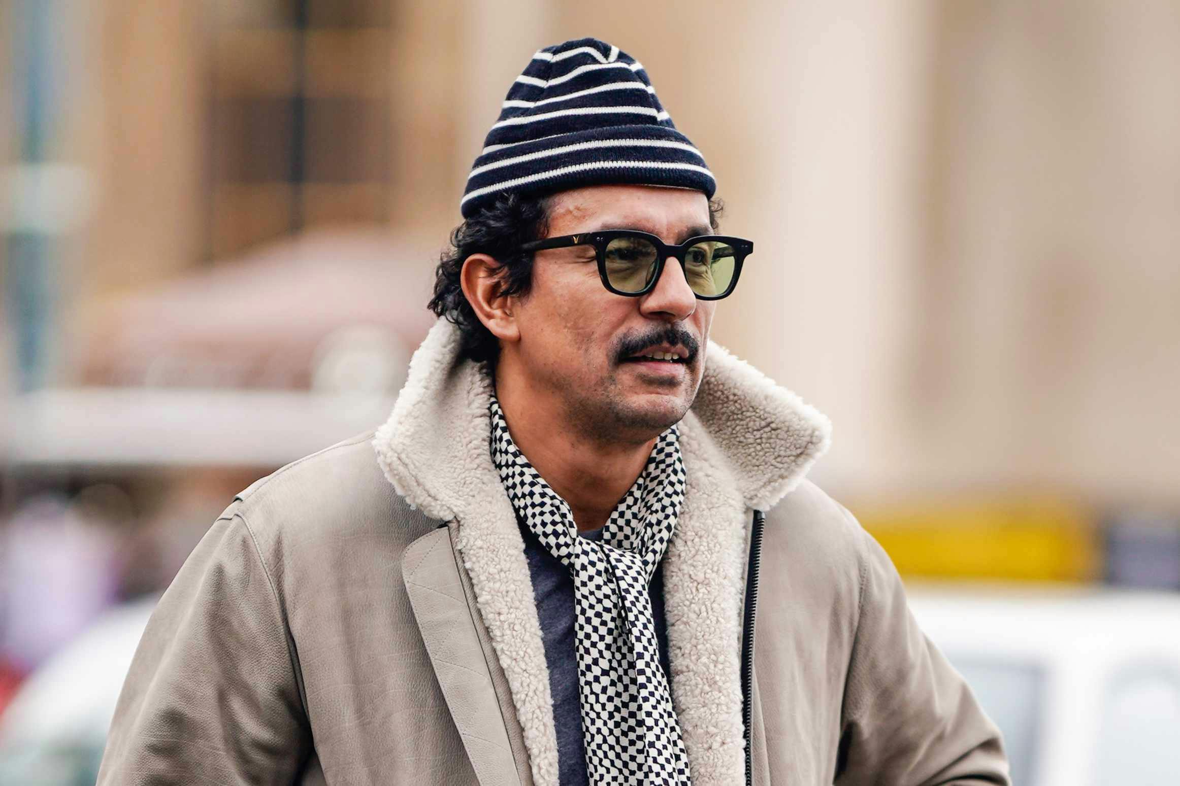 designer Haider Ackermann wears a grey jacket and striped hat