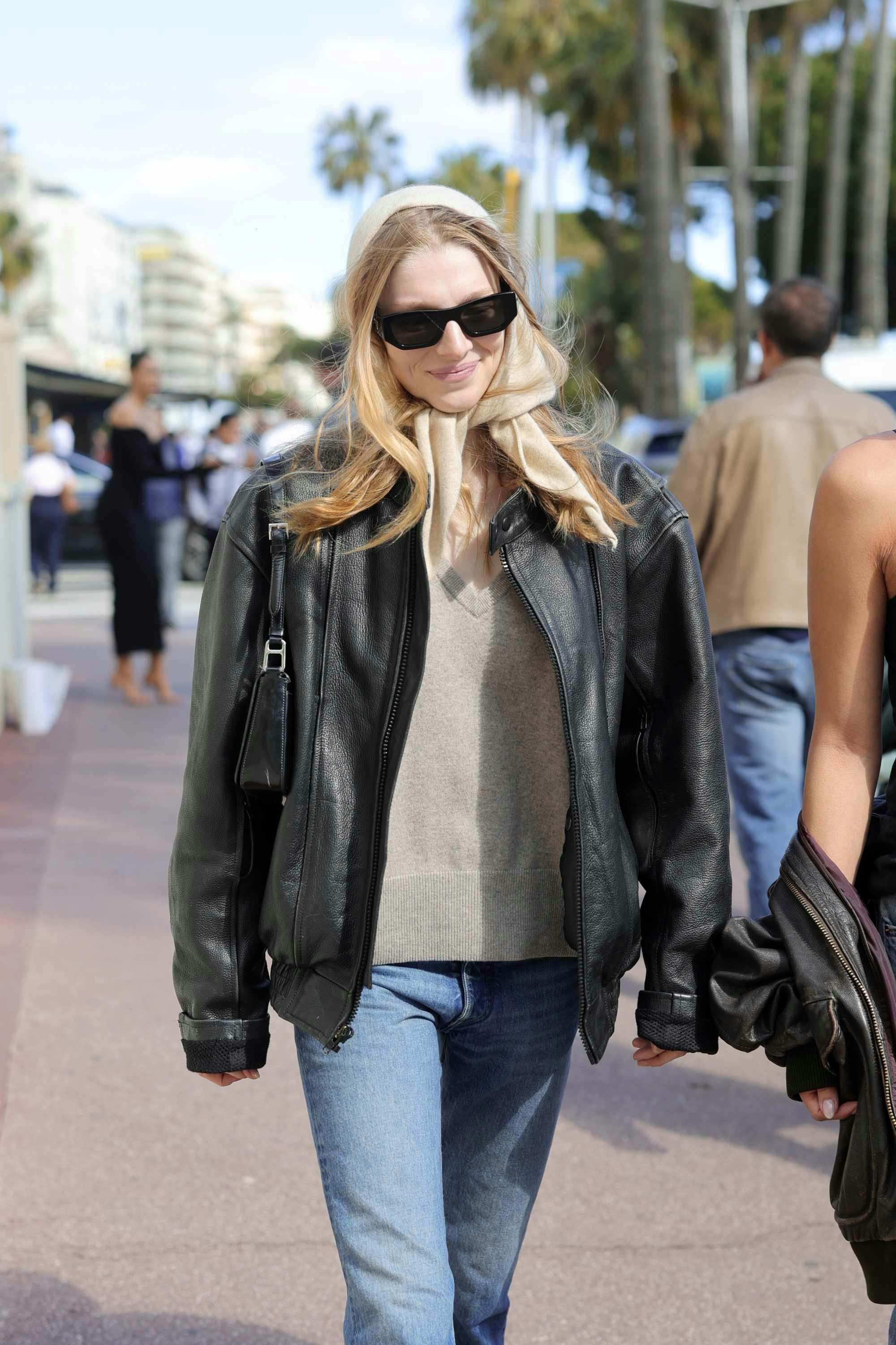 Hunter Schafer wears black Prada sunglasses with a brown leather jacket, beige sweater, blue jeans, and yellow headscarf at Cannes 2024