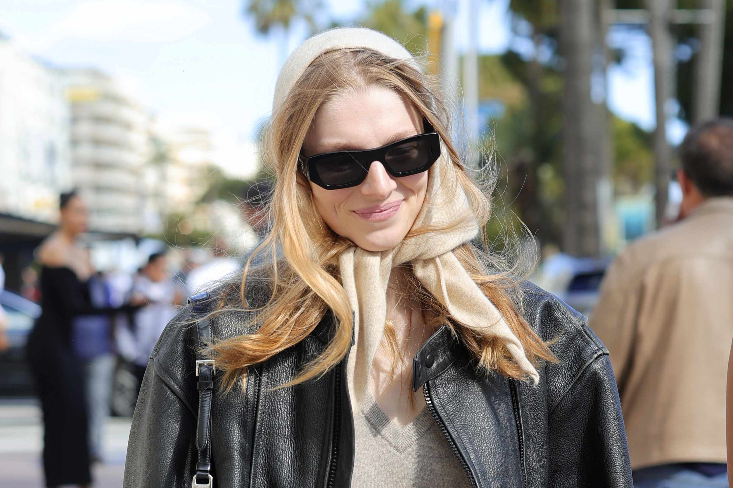 Hunter Schafer wears black Prada sunglasses with a brown leather jacket, beige sweater, blue jeans, and yellow headscarf at Cannes 2024