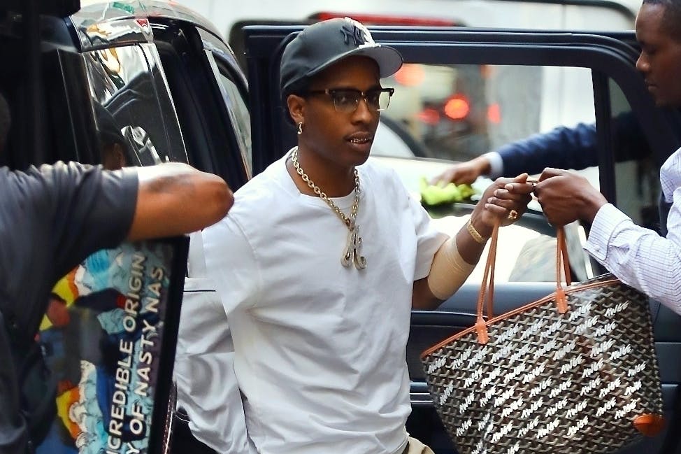 A$AP Rocky wears a white t-shirt, yankees hat, goyard awge bag, beige gold-studded pants, black leather shoes