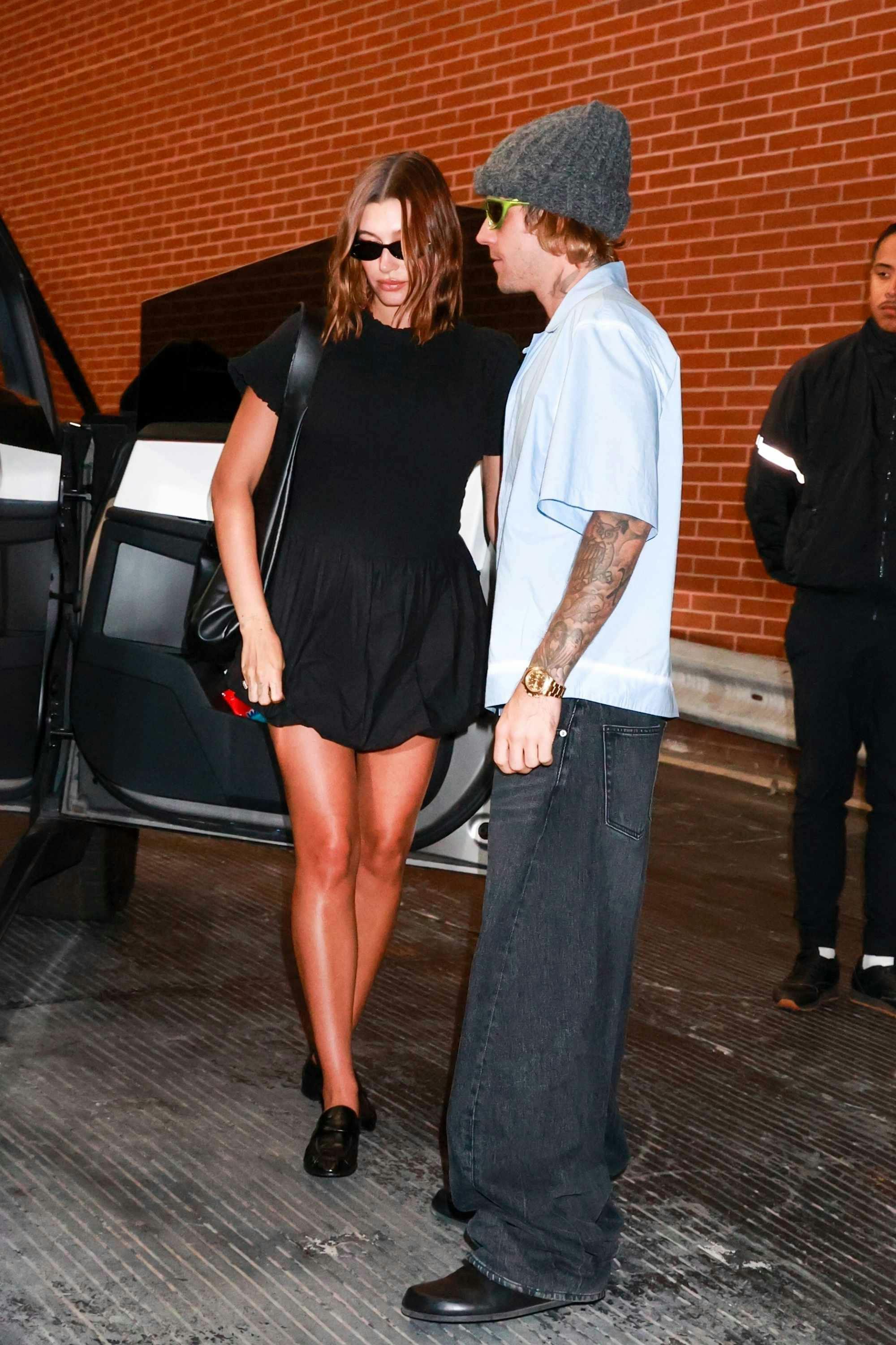 Justin Bieber wears grey beanie, technical sunglasses, baggy Balenciaga jeans & black clogs with Hailey Bieber in a black dress & sunglasses